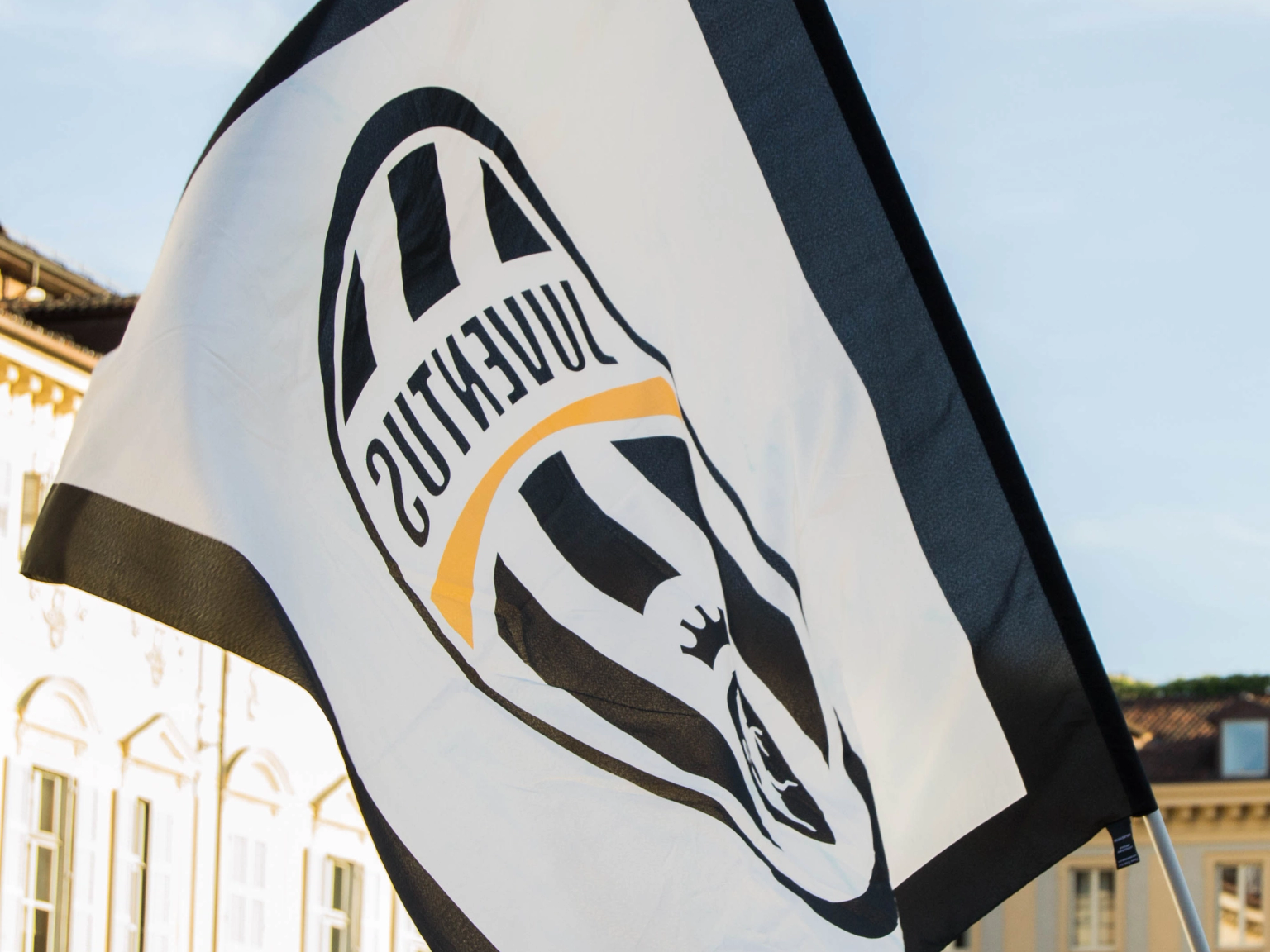 Eine Juventus-Flagge mit einem gespiegelten Logo weht vor historischen Gebäuden unter klarem Himmel.