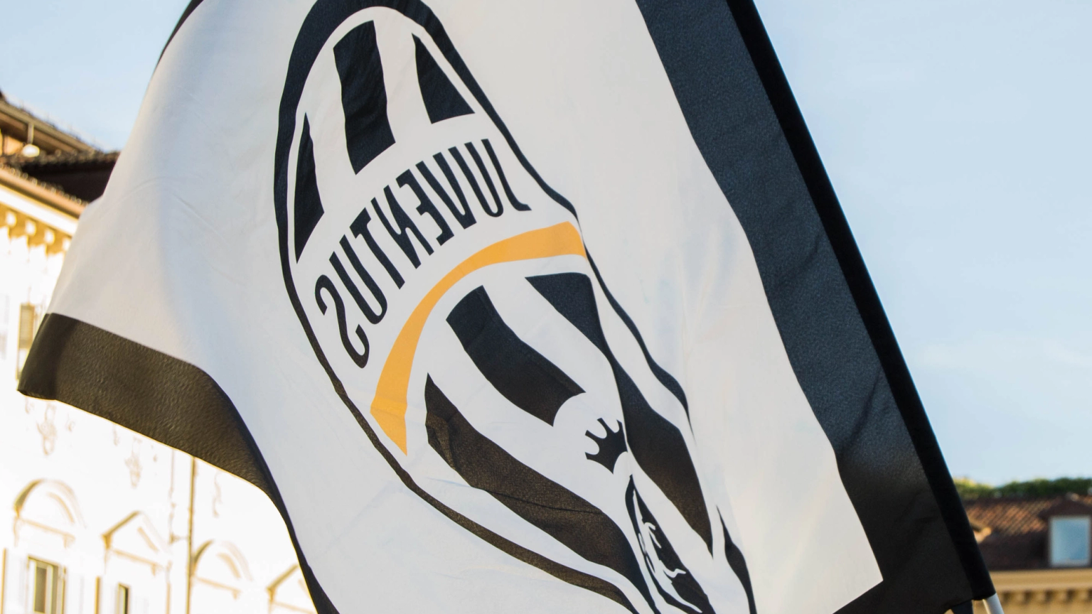 A Juventus flag with a mirrored logo waves in front of historic buildings under a clear sky.