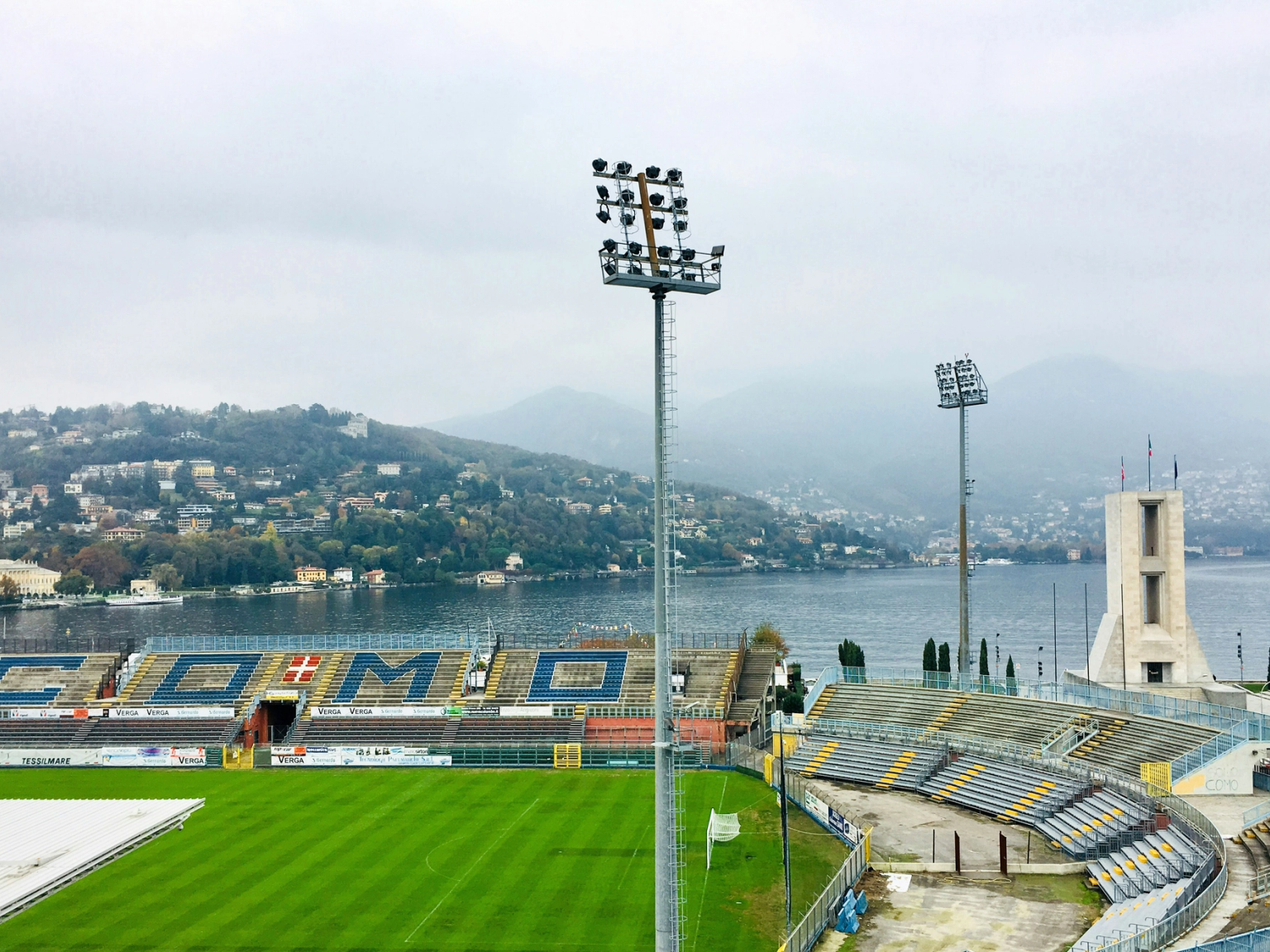An empty football stadium overlooking Lake Como, with mountains in the distance under a cloudy sky.