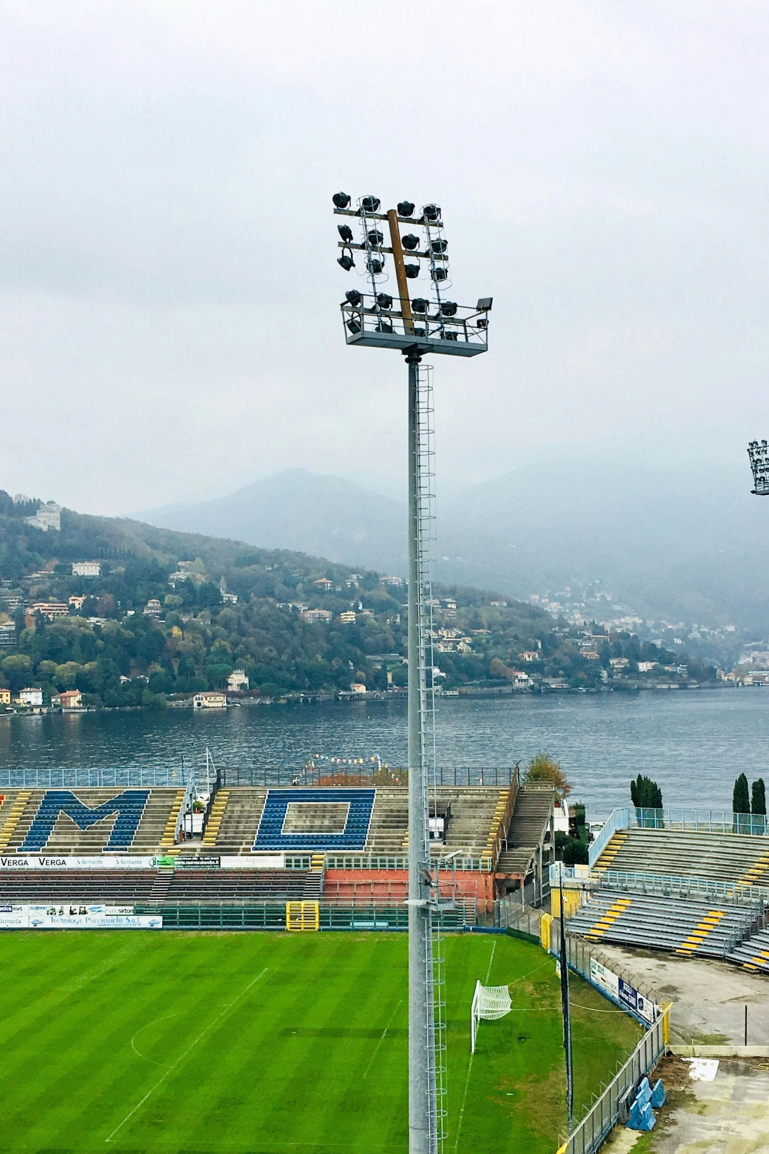 An empty football stadium overlooking Lake Como, with mountains in the distance under a cloudy sky.
