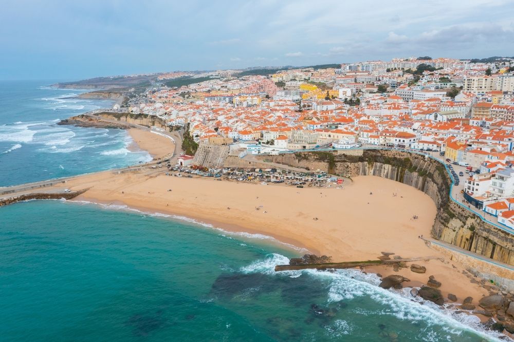 Flygfoto av en kuststad med röd-takade byggnader längs klippor som blickar ut över en sandstrand och turkost hav. Ericeira, Portugal