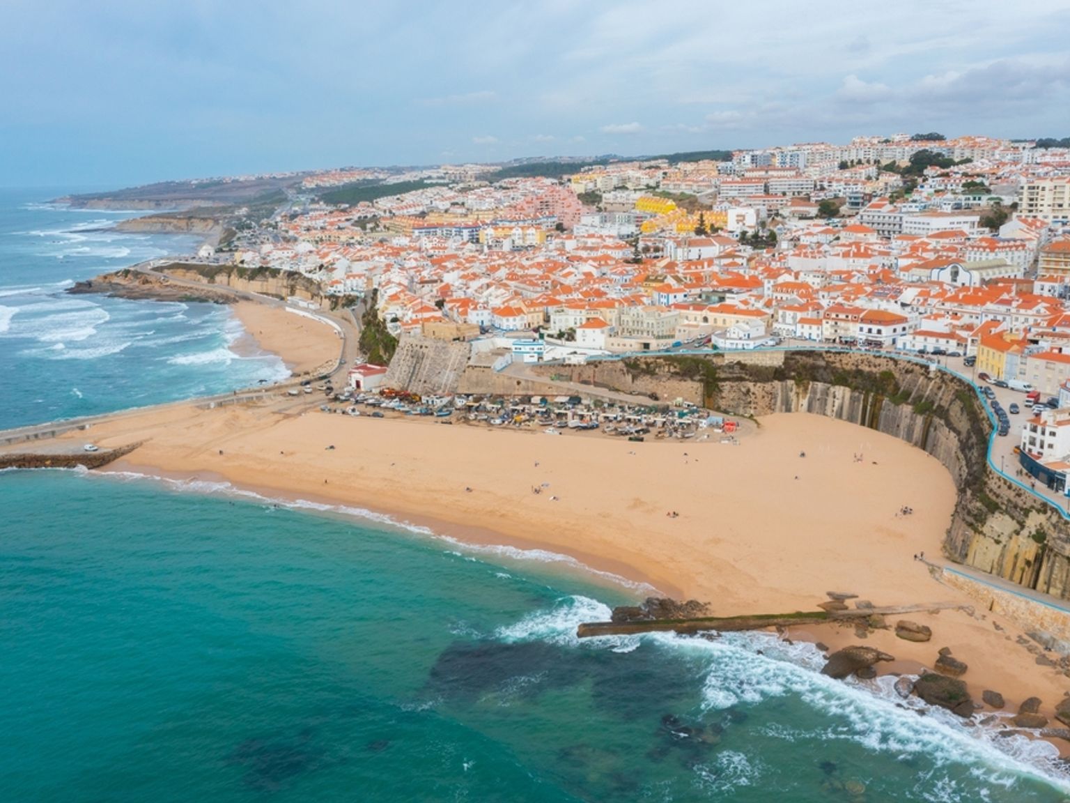 Flygfoto av en kuststad med röd-takade byggnader längs klippor som blickar ut över en sandstrand och turkost hav. Ericeira, Portugal