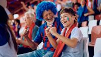 An older man in a blue wig and a young boy with face paint cheer happily at a sports game.