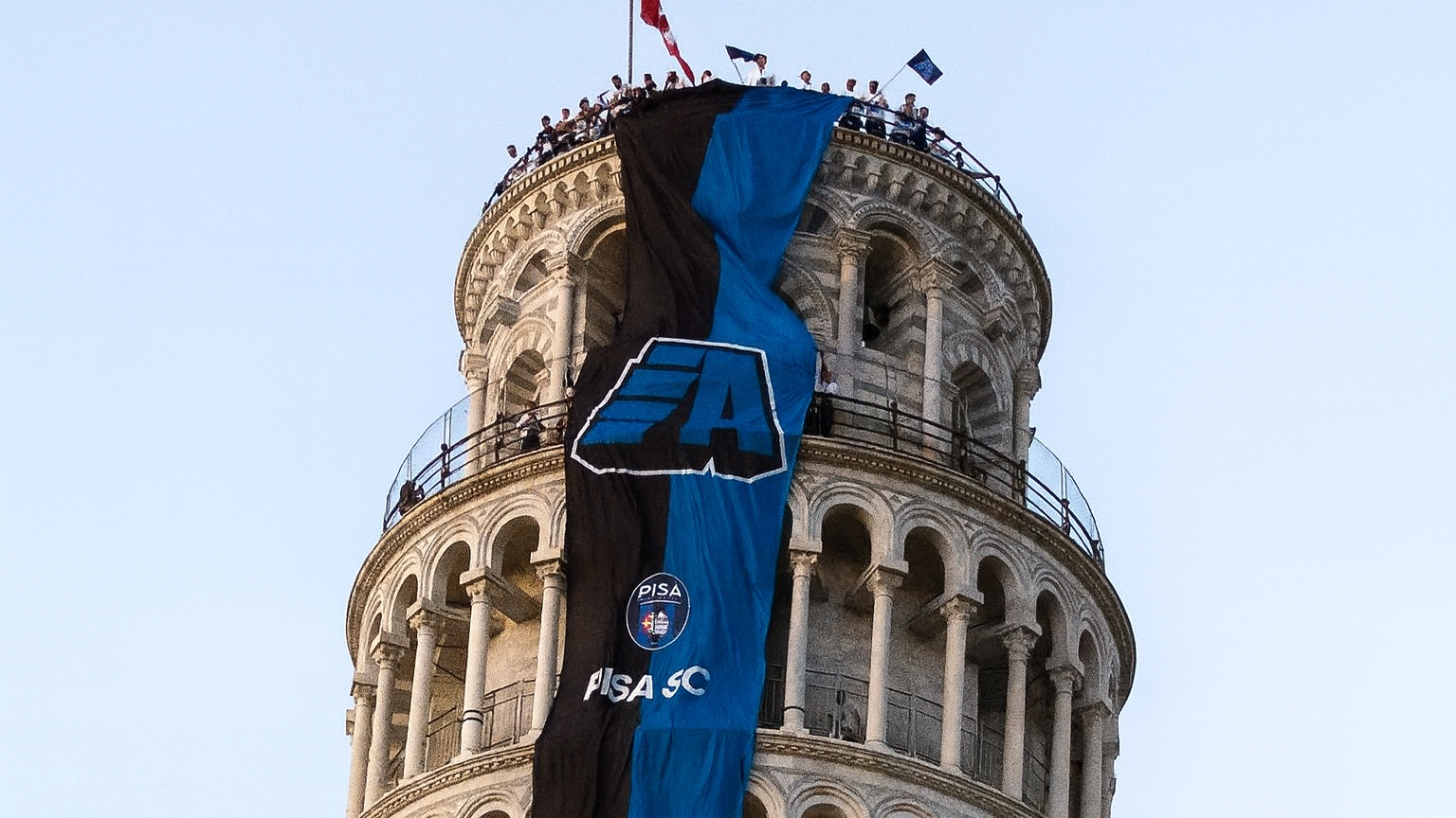 Toppen av Lutande tornet i Pisa med en stor svart och blå Pisa SC-banner som hänger ner, och människor på observationsdäcket.