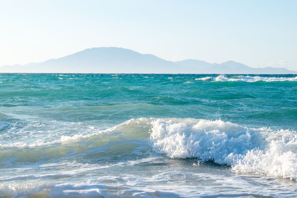 Turkosa vågor bryter på en strand med dimmiga berg i fjärran. Tsilivi, Zakhyntos