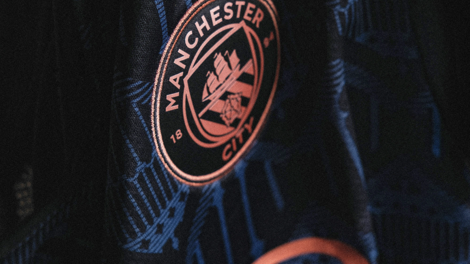 A close-up of a dark jersey featuring a metallic copper Manchester City FC crest and a subtle blue geometric pattern.