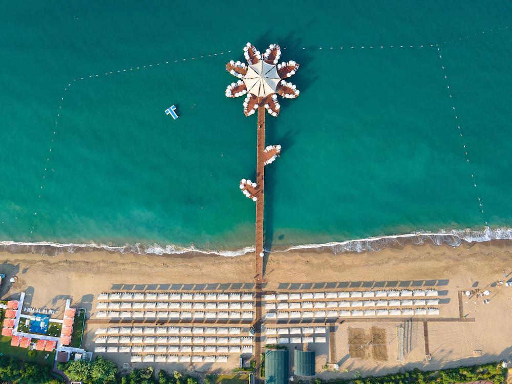 Aerial view of a sandy beach with a flower-shaped pier extending over turquoise water, flanked by rows of white beach cabanas. Sueno Hotels Beach Side