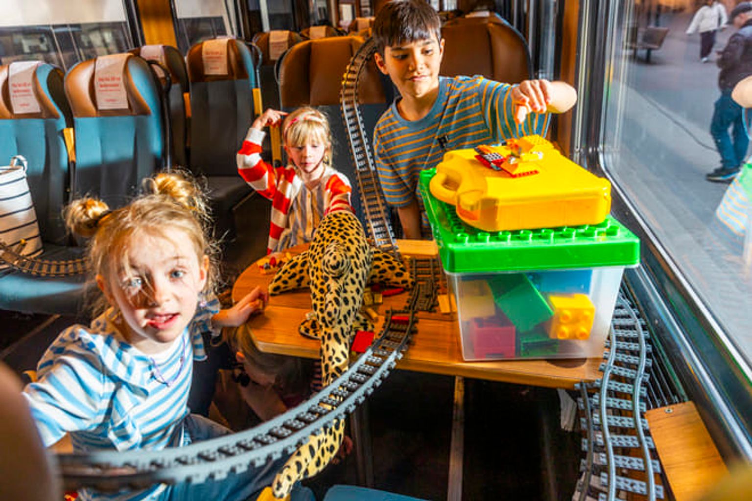 Tre barn leker med leksakstågräls och byggklossar i en tågvagn.