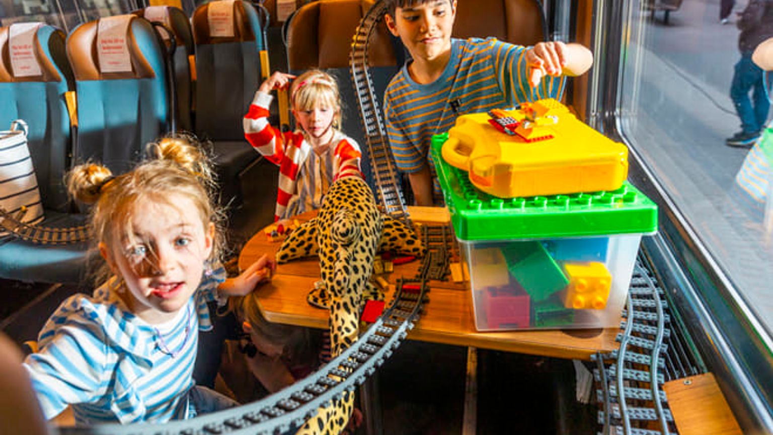 Tre barn leker med leksakstågräls och byggklossar i en tågvagn.