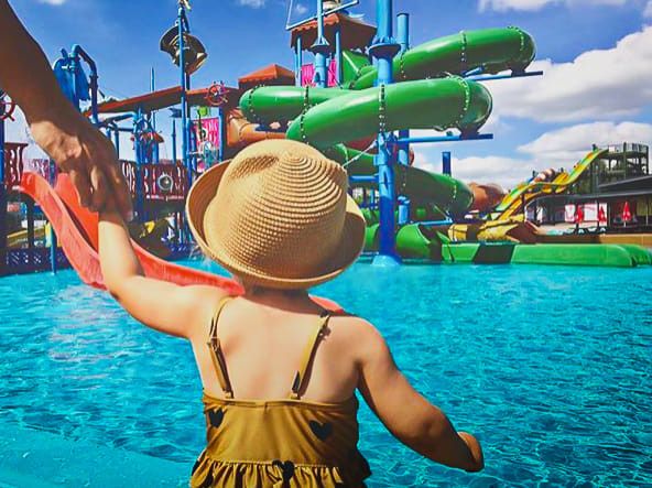 Et barn med stråhat holder en voksens hånd og kigger ud over et vandland med farverige rutsjebaner.