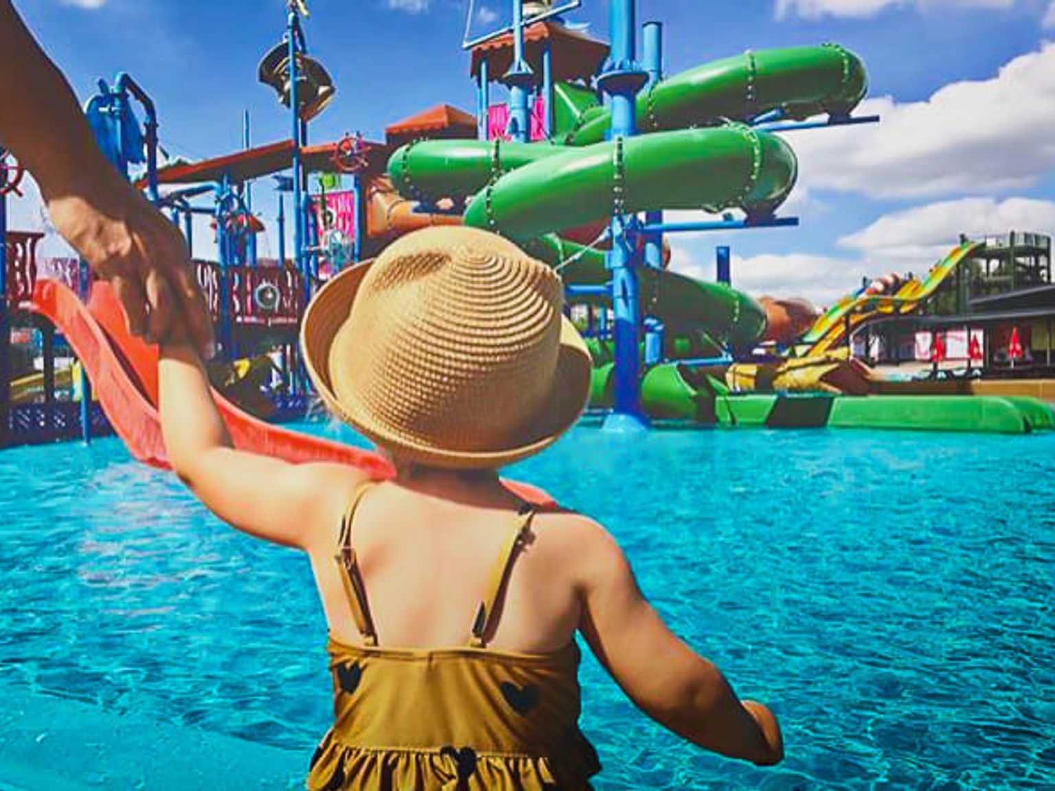 Et barn med stråhat holder en voksens hånd og kigger ud over et vandland med farverige rutsjebaner.