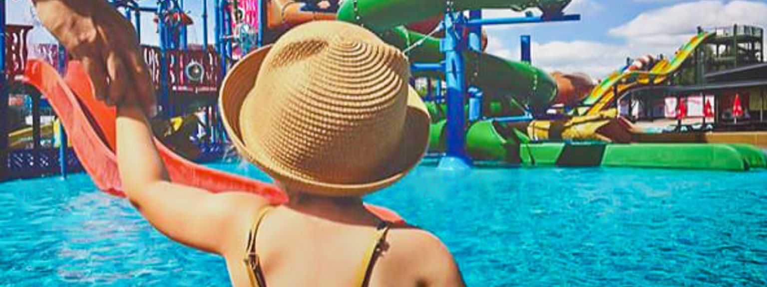 Et barn med stråhat holder en voksens hånd og kigger ud over et vandland med farverige rutsjebaner.