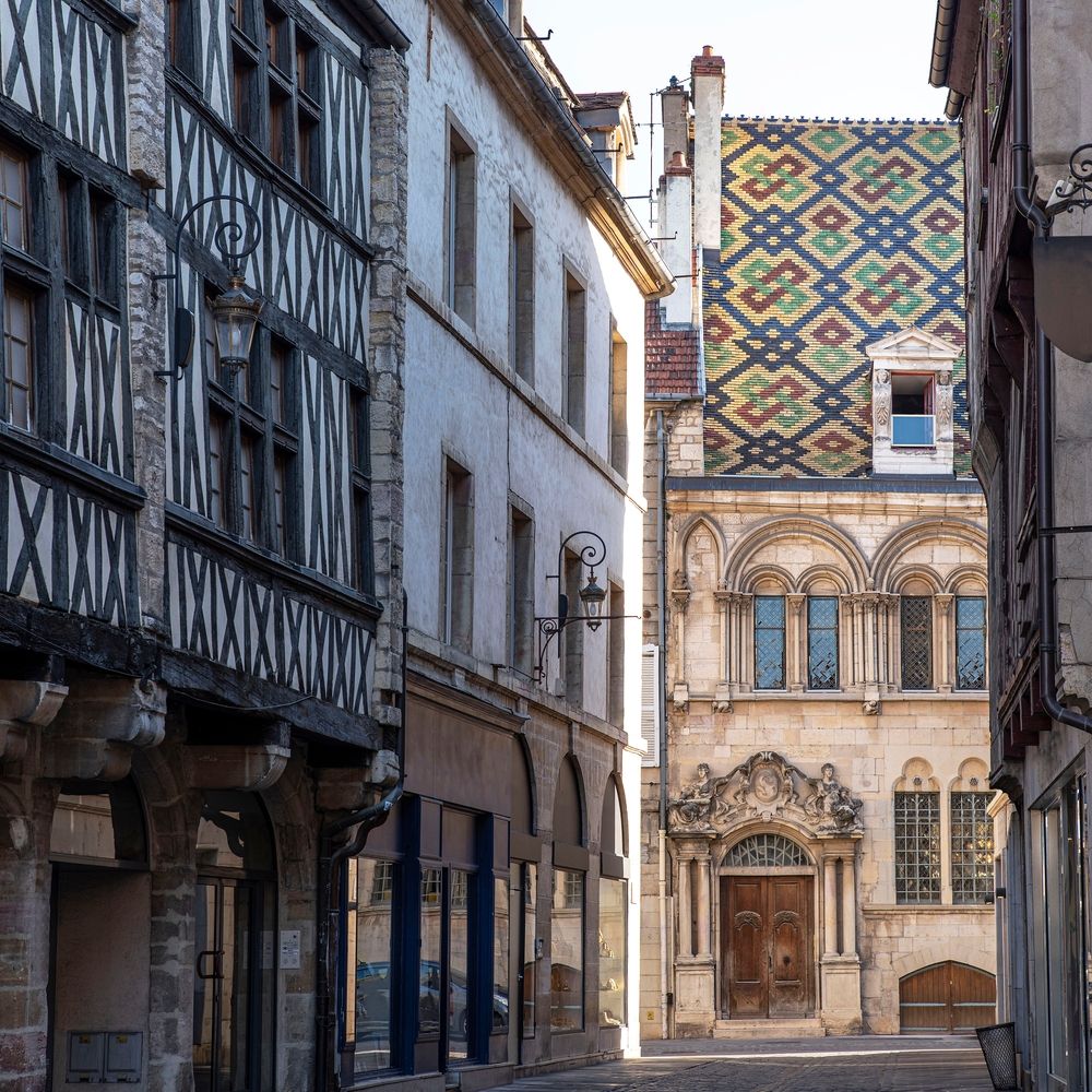 A narrow street lined with half-timbered buildings, leading to a historic building with an ornate entrance and a colorful patterned roof. Dijon, France