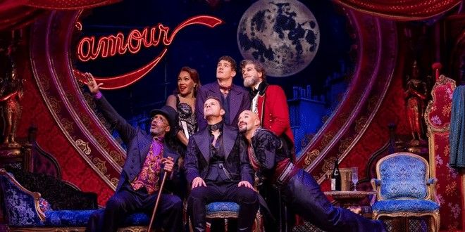 Sieben Künstler in aufwendigen Kostümen posieren auf einer Theaterbühne mit einem roten Herz, einem Neonschild mit der Aufschrift „l'amour“ und einem großen Mond.