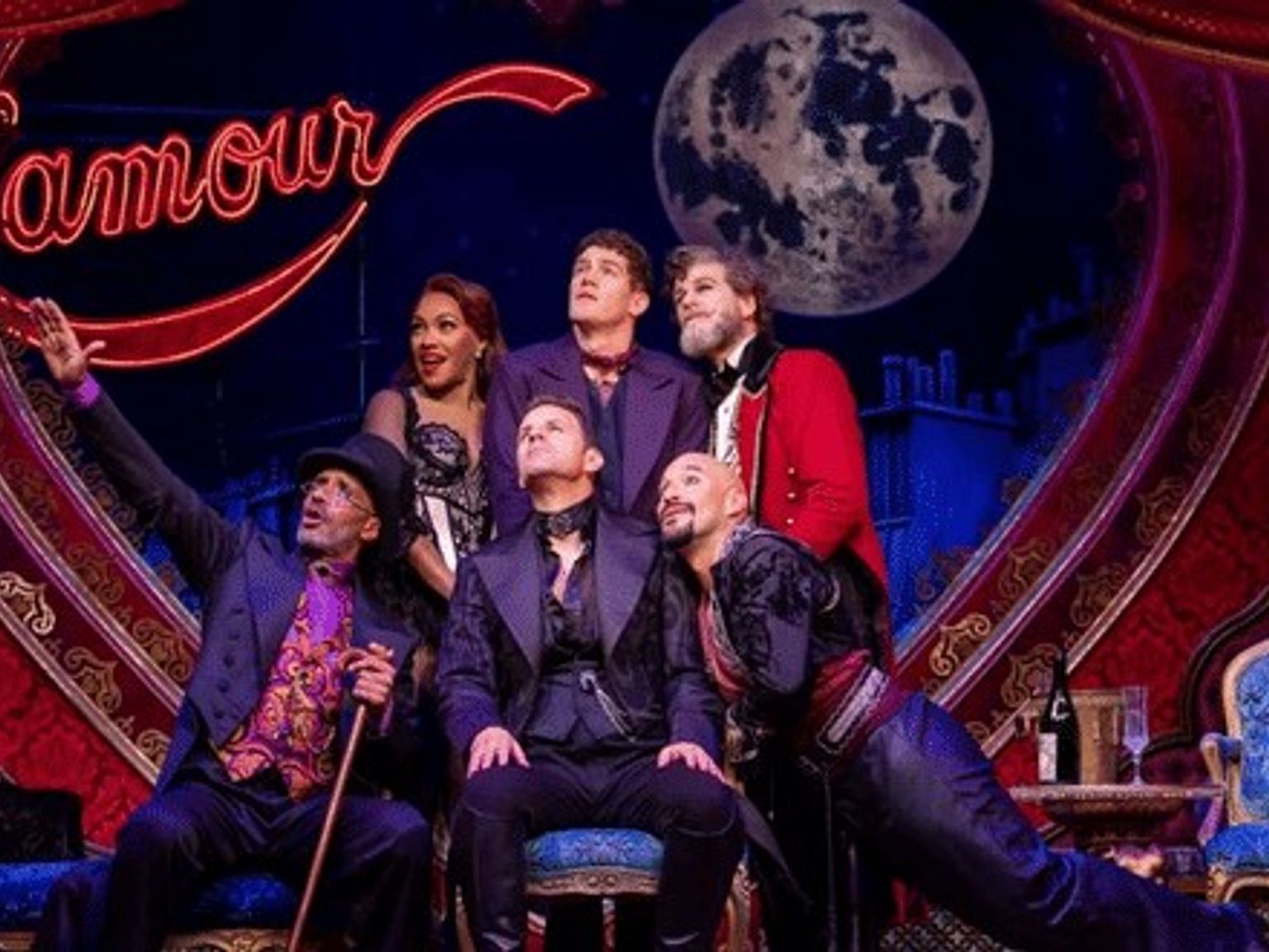 Sieben Künstler in aufwendigen Kostümen posieren auf einer Theaterbühne mit einem roten Herz, einem Neonschild mit der Aufschrift „l'amour“ und einem großen Mond.