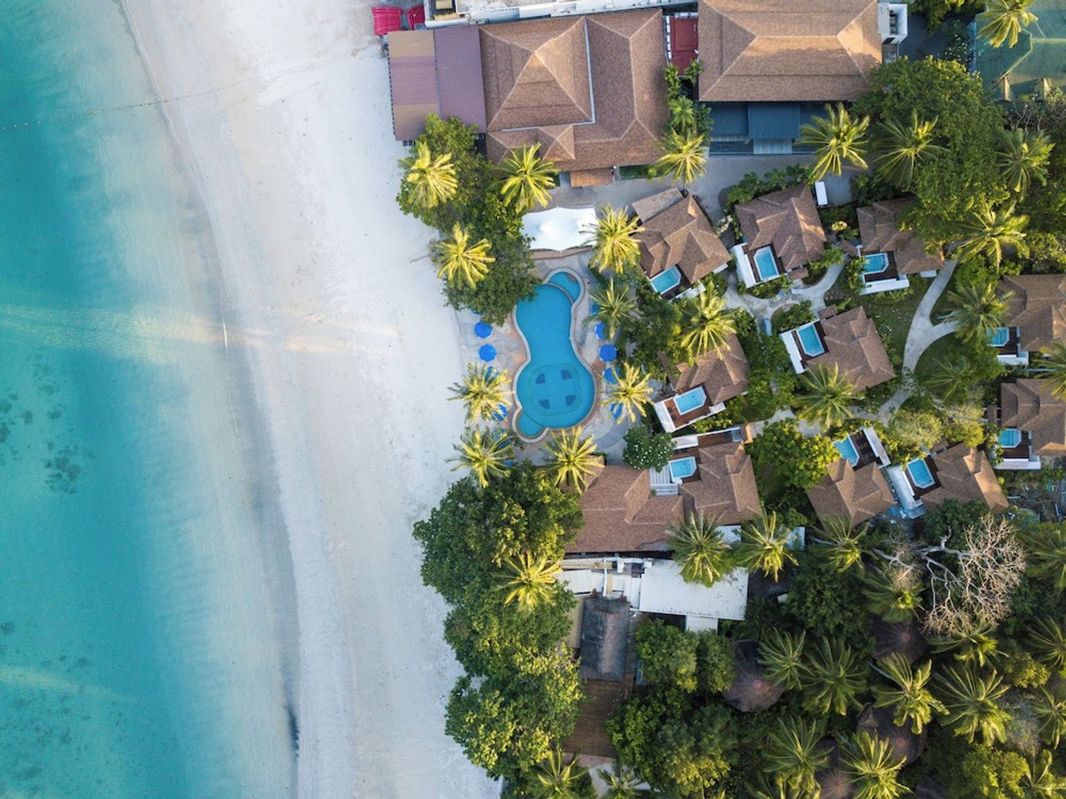 Vista aérea de un complejo turístico tropical con bungalós con piscina privada, una gran piscina central, palmeras, una playa de arena blanca y un océano turquesa.
