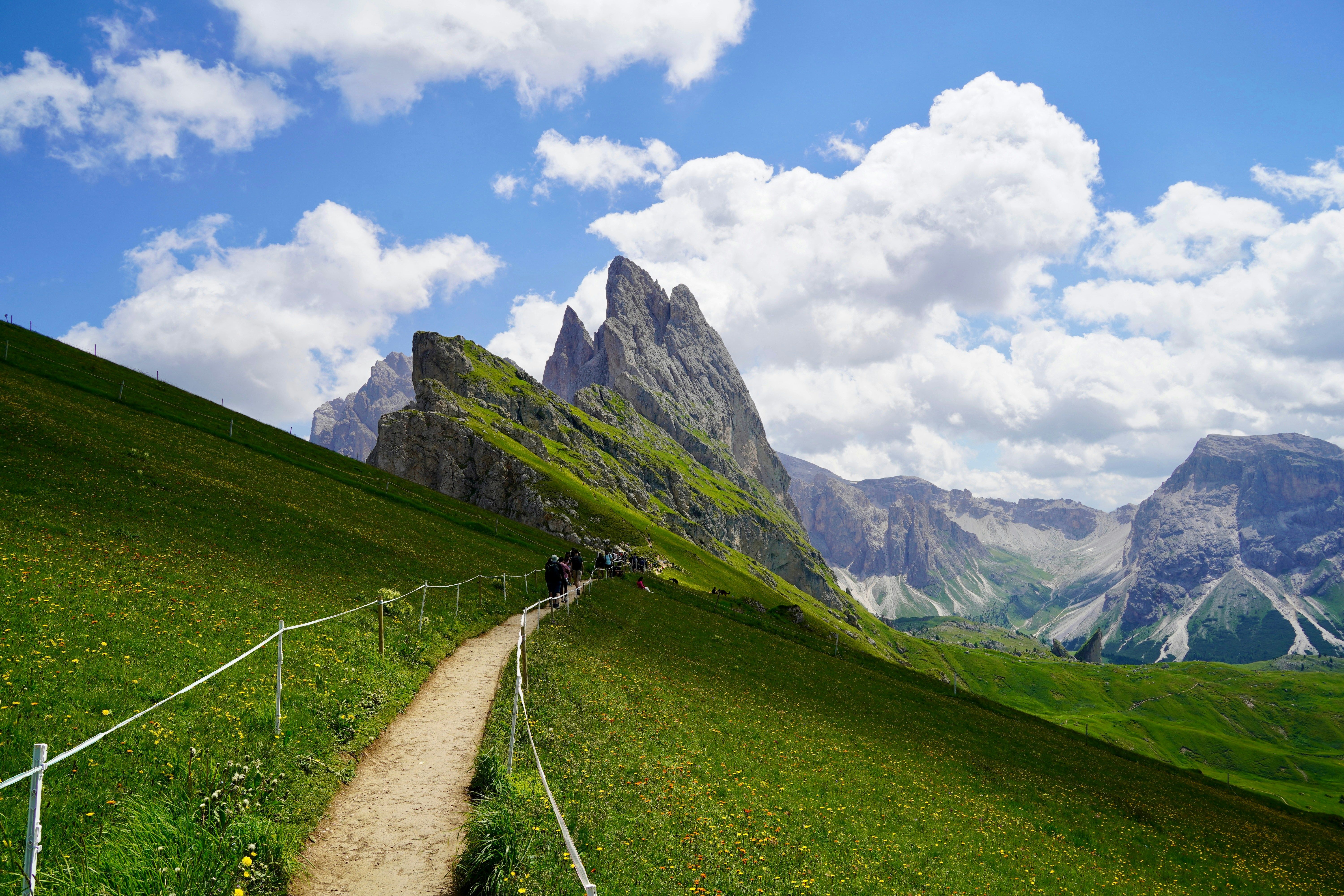 En grussti slynger seg gjennom en grønn fjelleng mot taggete topper under en blå himmel med skyer. Seceda, Ortisei, Italia
