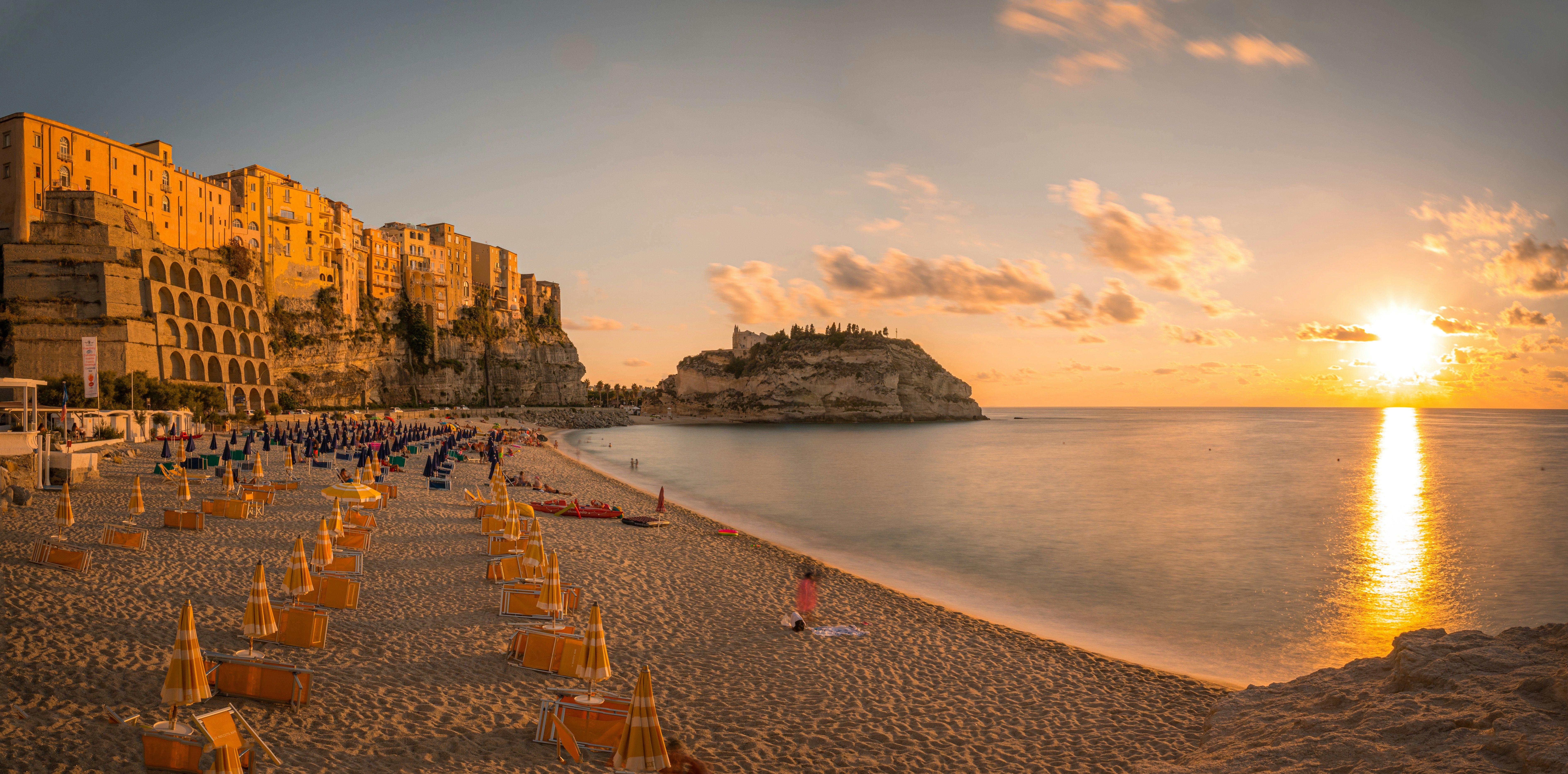 Gylden solnedgang over en sandstrand med parasoller, en kystby på klipper og en ø med bygninger. Tropea, Calabria