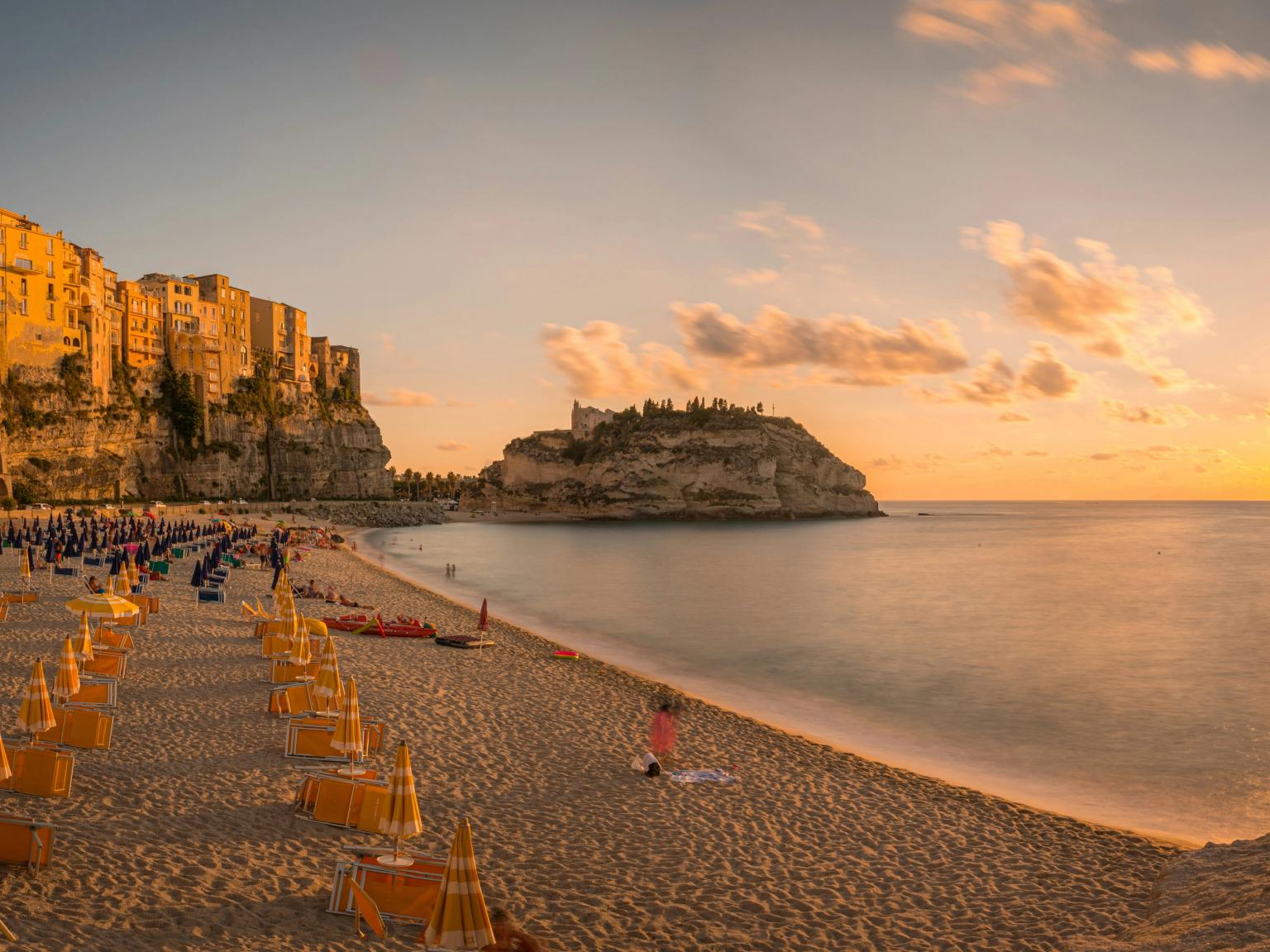 Tramonto dorato su una spiaggia sabbiosa costeggiata da ombrelloni, una città costiera arroccata sulle scogliere e un’isola con edifici. Tropea, Calabria