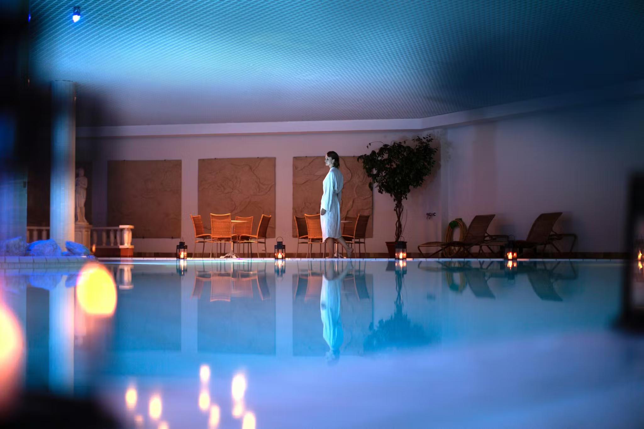 A woman in a white bathrobe stands beside a glowing blue indoor pool, reflecting atmospheric candlelight.