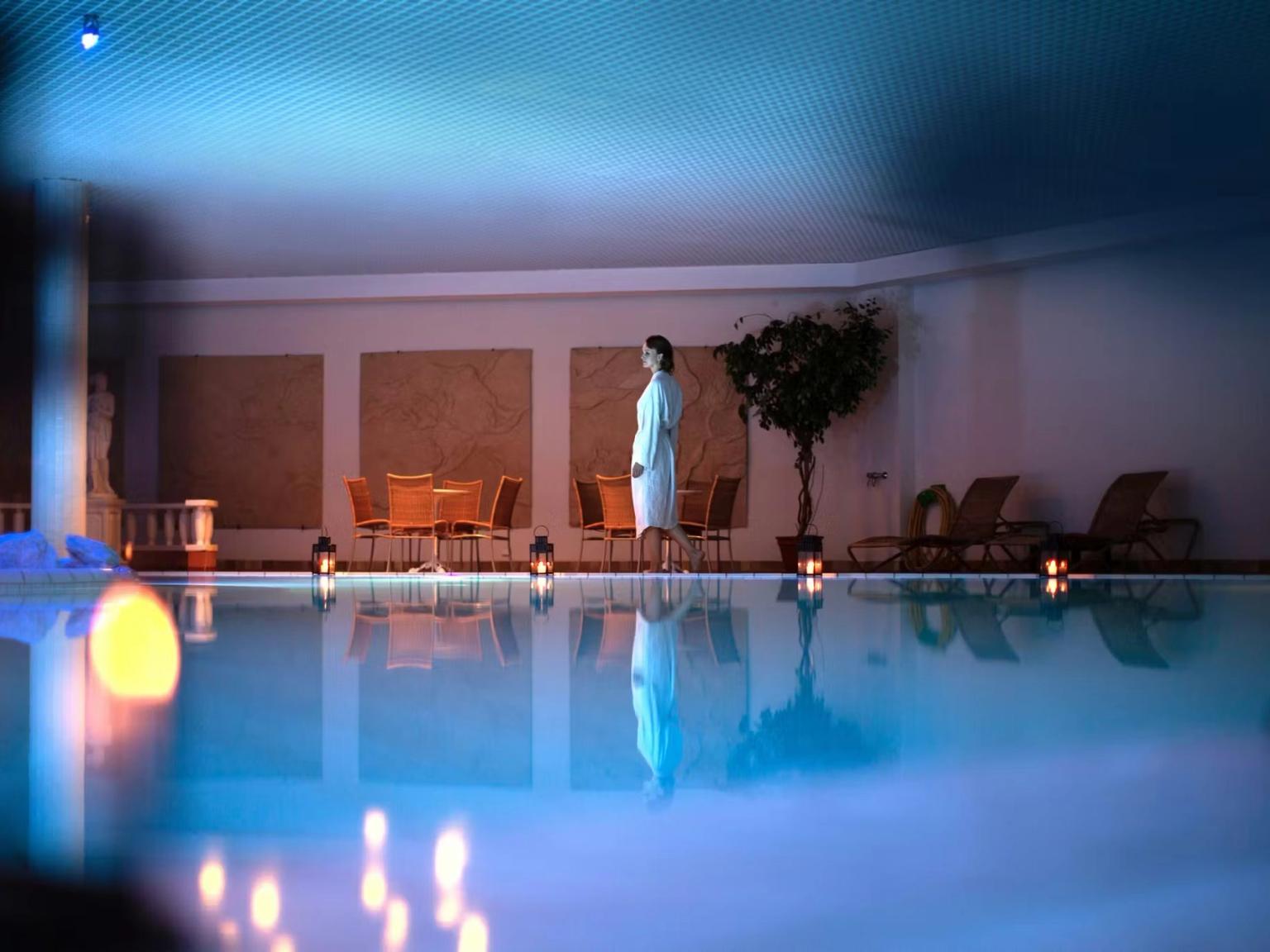 A woman in a white bathrobe stands beside a glowing blue indoor pool, reflecting atmospheric candlelight.