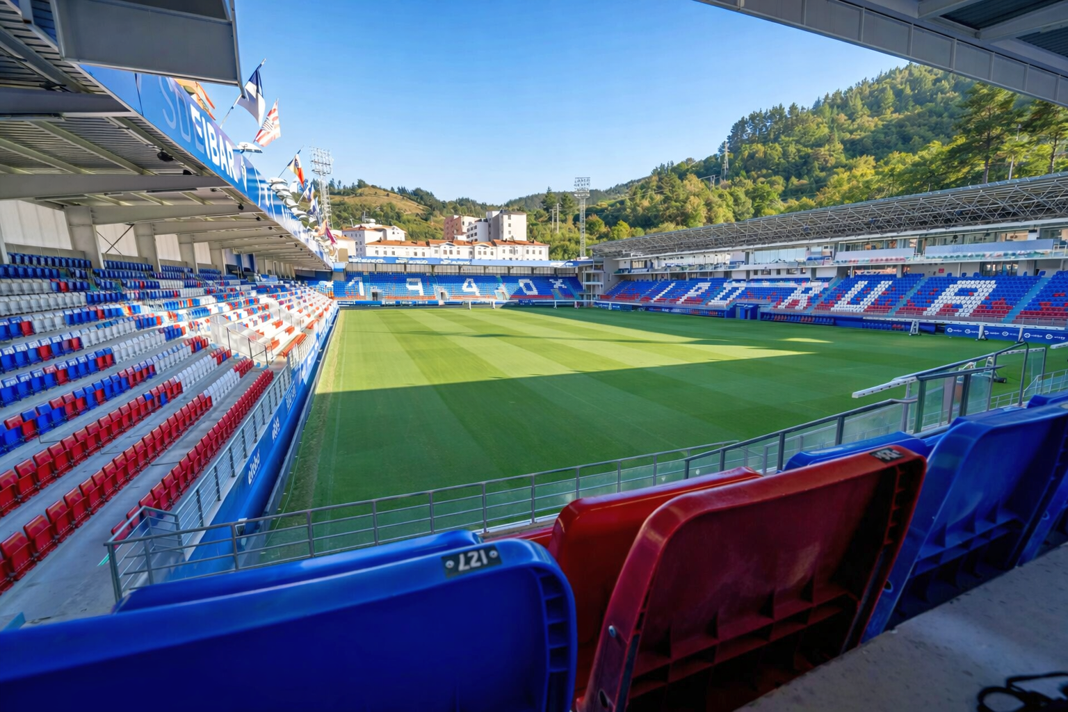 Empty football stadium with a green pitch, red and blue seats, and hills in the background under a blue sky.