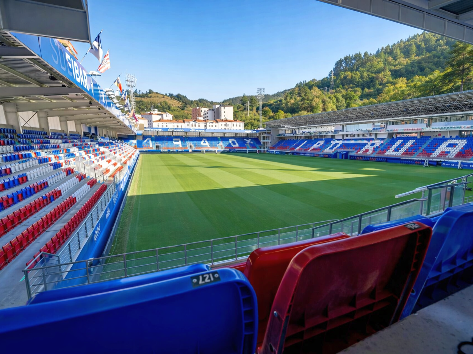 Empty football stadium with a green pitch, red and blue seats, and hills in the background under a blue sky.