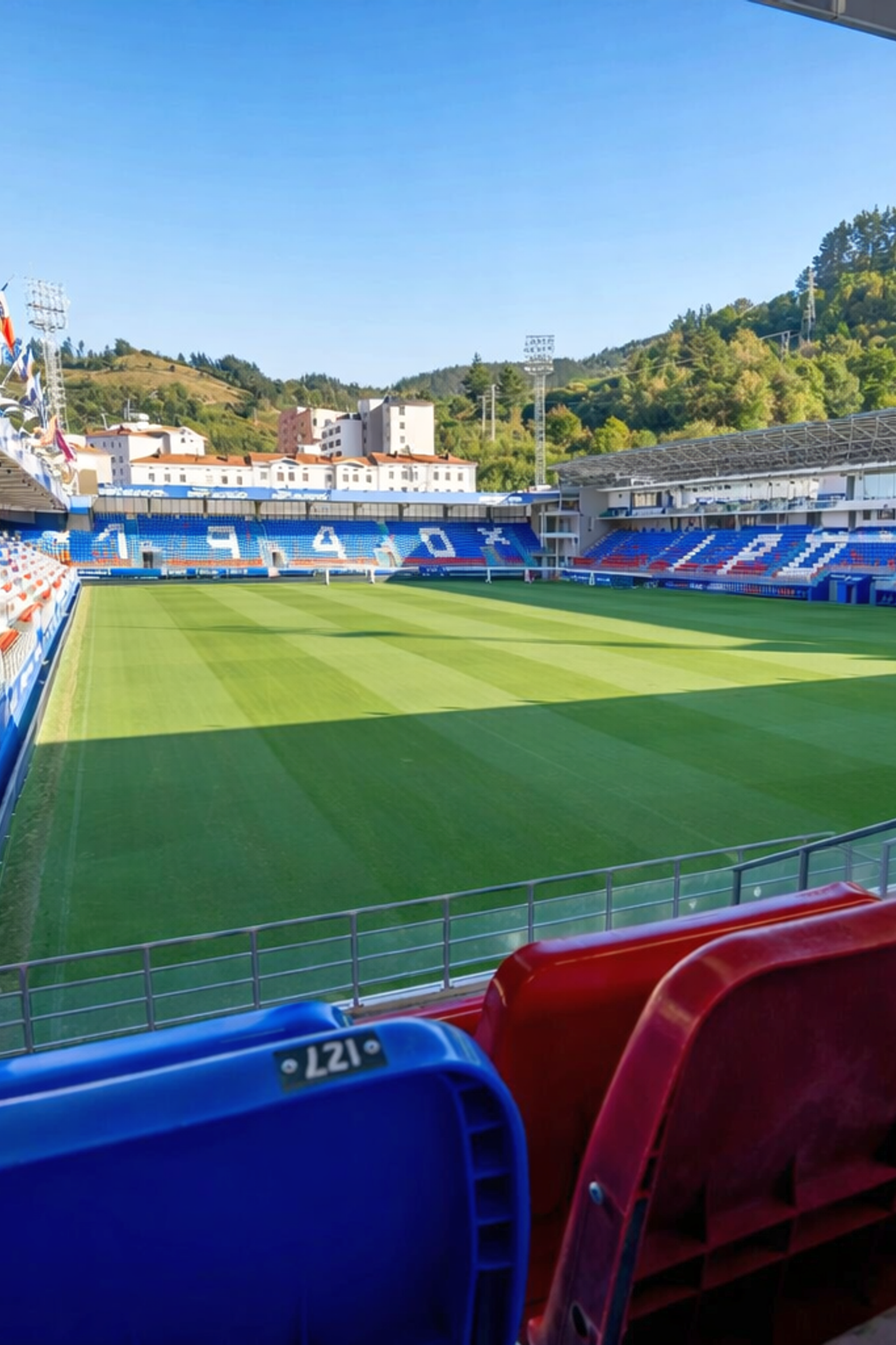 Empty football stadium with a green pitch, red and blue seats, and hills in the background under a blue sky.