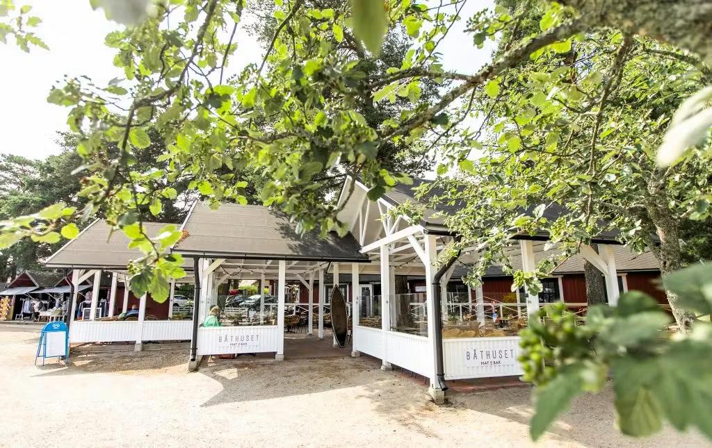 En rød og hvid bygning med en udendørs terrasse omgivet af grønne træer på en solrig dag.