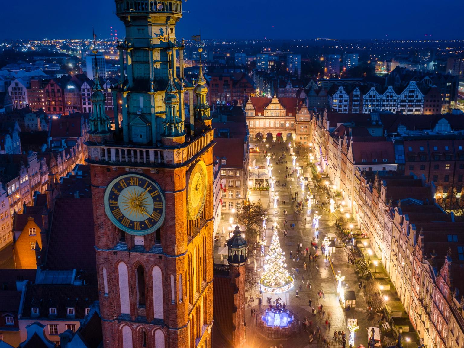 Nattlig flygvy över Gdansk med ett framträdande klocktorn, en stor upplyst julgran och festliga ljus.