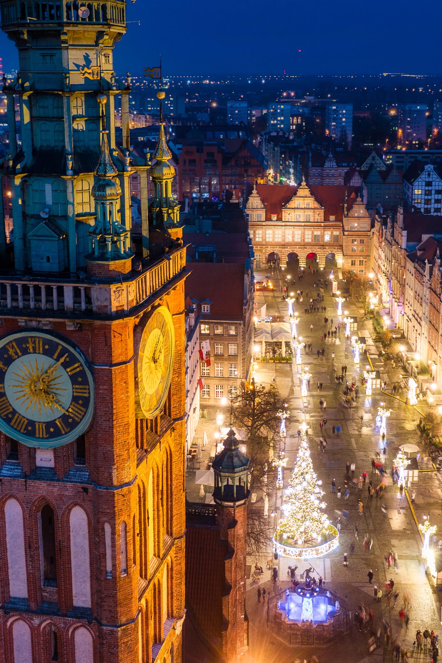 Nattlig flygvy över Gdansk med ett framträdande klocktorn, en stor upplyst julgran och festliga ljus.