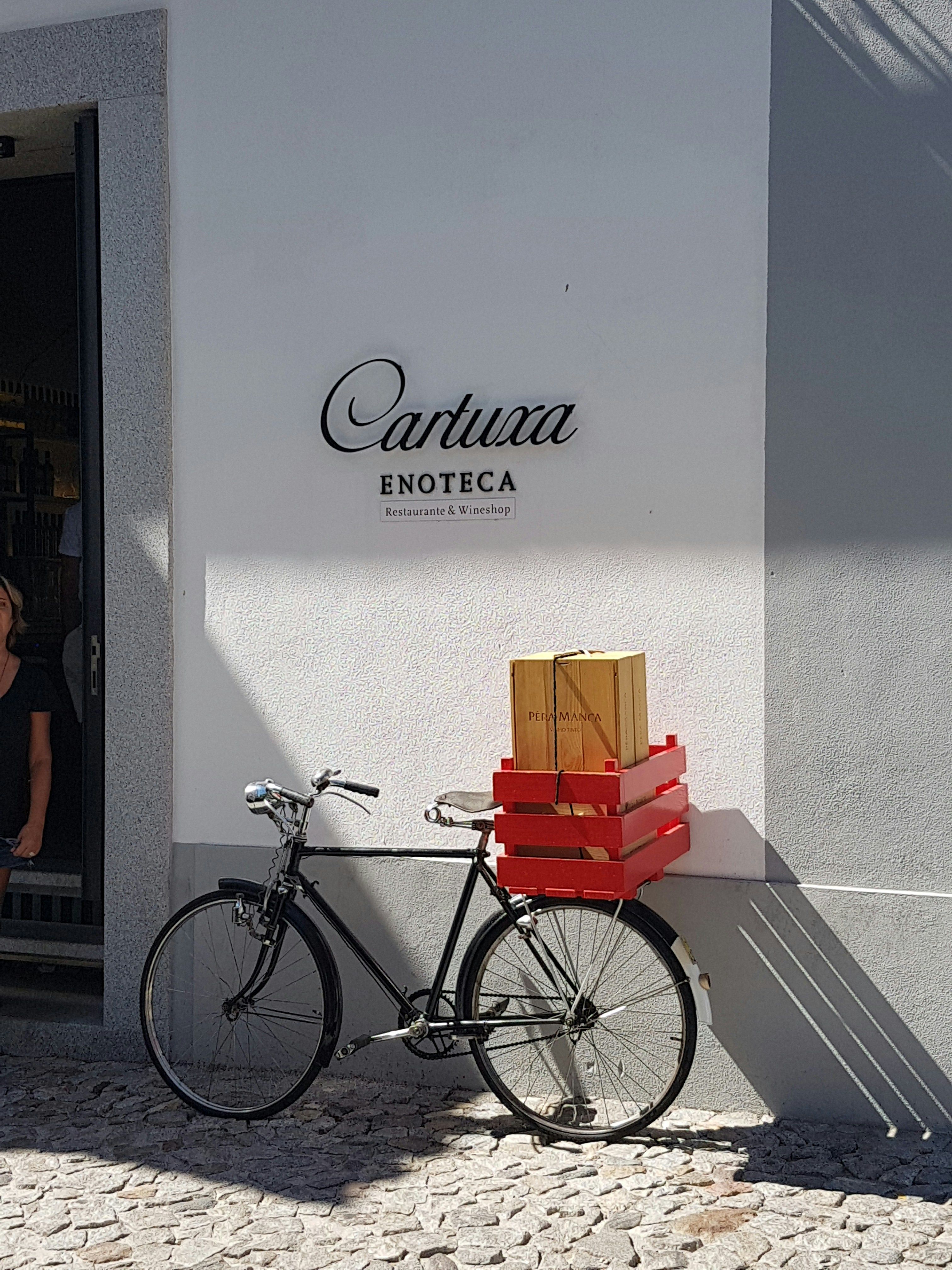 En svart cykel med röda backar och en Pêra Manca-vinlåda står parkerad mot en vit vägg med skylten Cartuxa Enoteca.