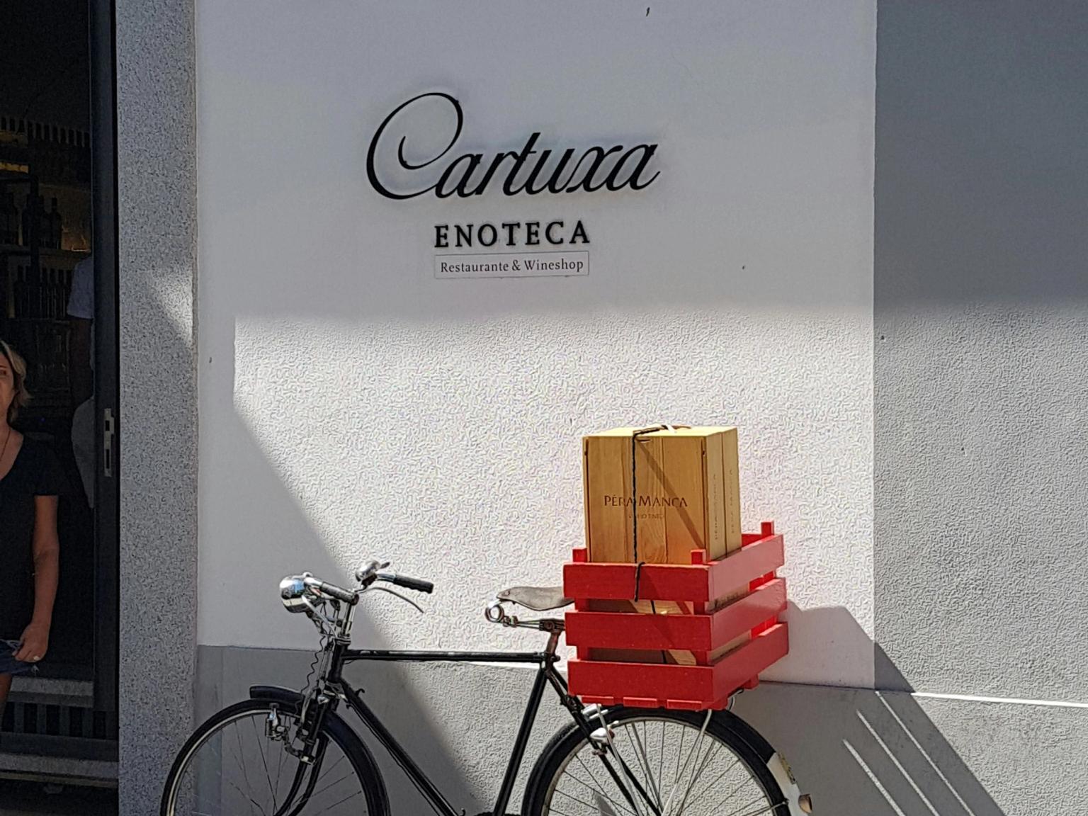 En svart cykel med röda backar och en Pêra Manca-vinlåda står parkerad mot en vit vägg med skylten Cartuxa Enoteca.