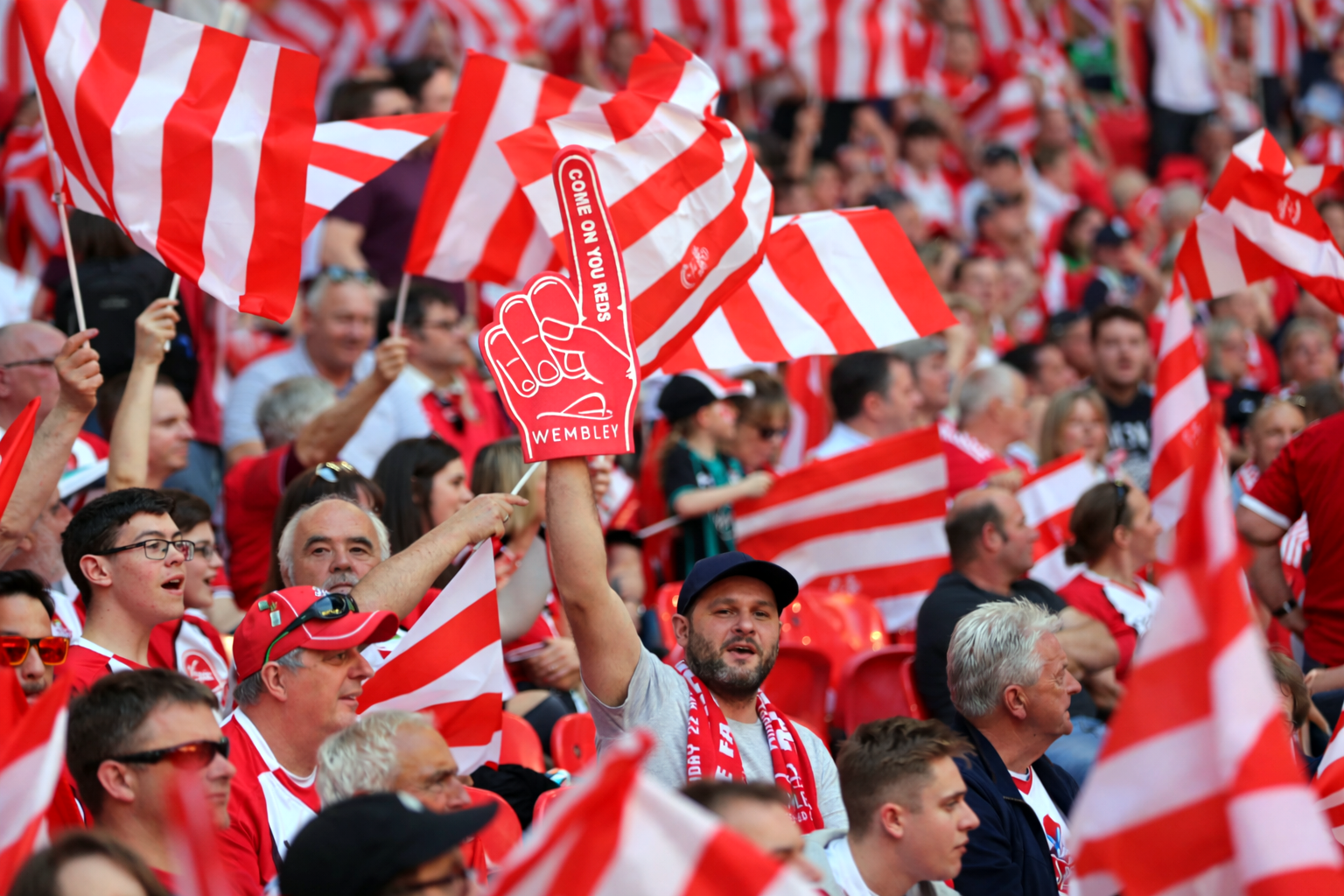 A stadium crowd, many holding red and white flags. One fan raises a red foam finger displaying 'COME ON YOU REDS' and 'WEMBLEY'.