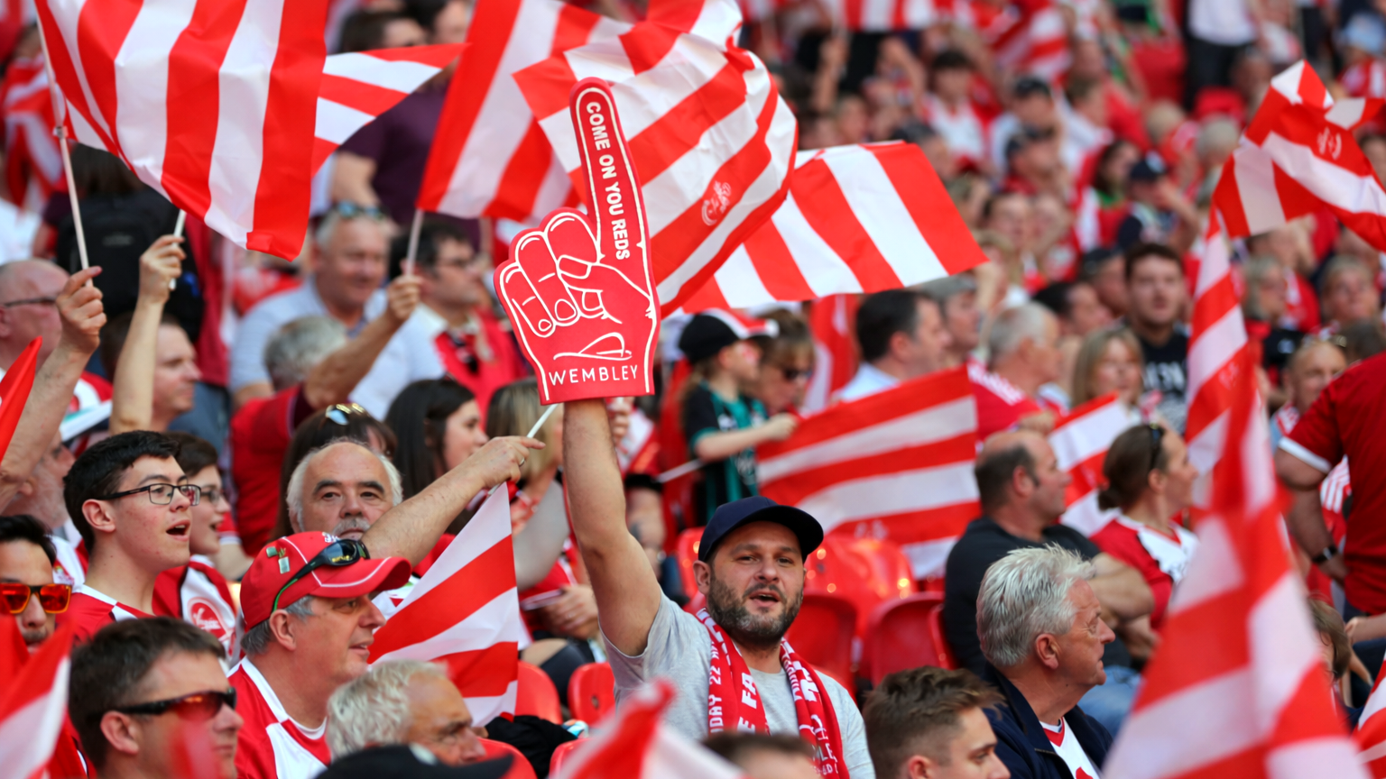 A stadium crowd, many holding red and white flags. One fan raises a red foam finger displaying 'COME ON YOU REDS' and 'WEMBLEY'.