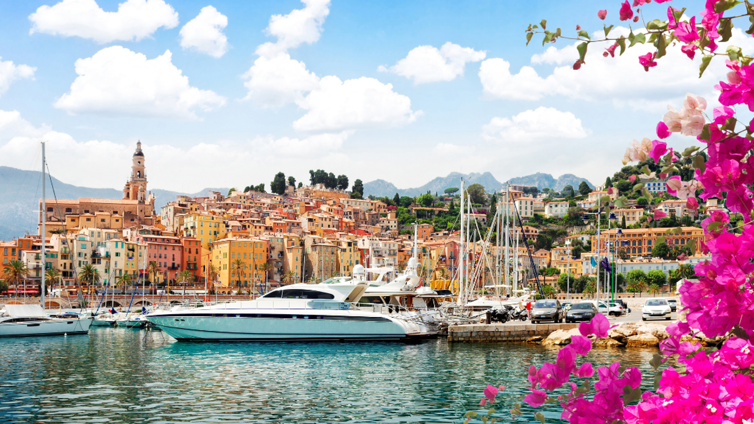 Livlig medelhavshamn på Franska rivieran med färgglada byggnader, lyxyachter och rosa blommor.