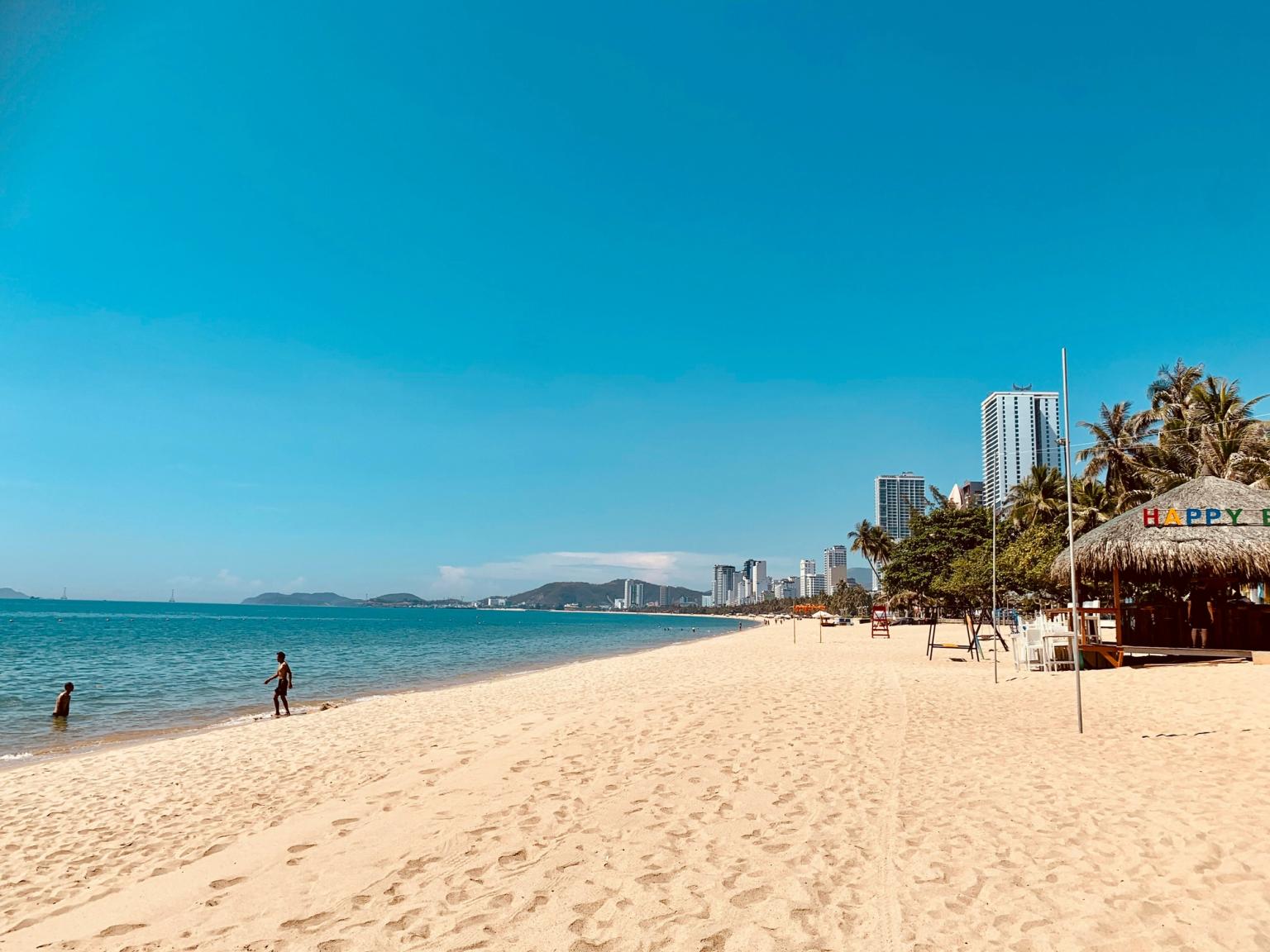 En bred sandstrand med blått vatten, en avlägsen stadssilhuett och en halmtäckt "Happy Bar"-hydda.