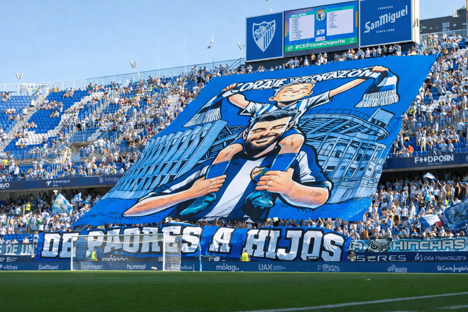 A large blue and white banner with a cartoon of a father carrying his son and the text "DE PADRES A HIJOS" is unfurled by fans in a stadium.