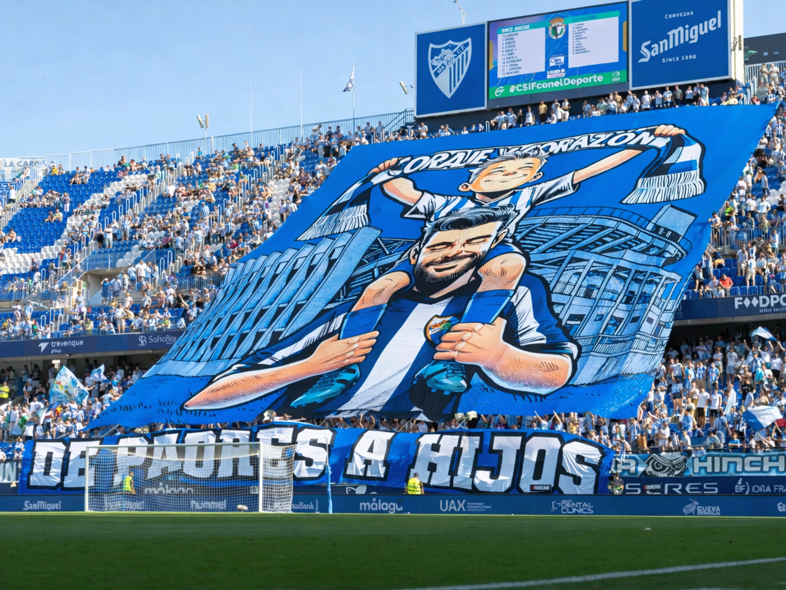 A large blue and white banner with a cartoon of a father carrying his son and the text "DE PADRES A HIJOS" is unfurled by fans in a stadium.