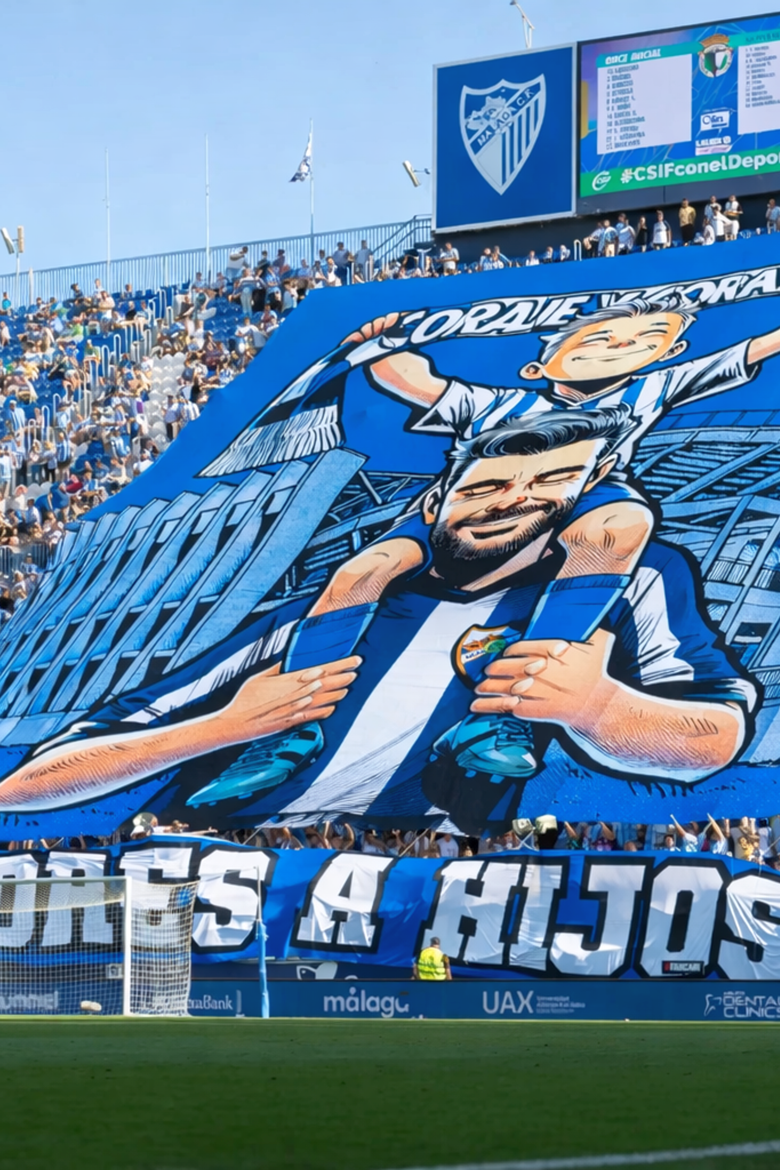 A large blue and white banner with a cartoon of a father carrying his son and the text "DE PADRES A HIJOS" is unfurled by fans in a stadium.
