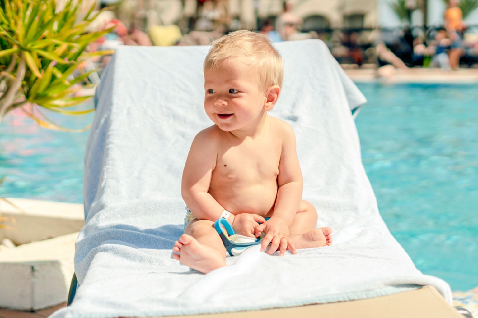 En smilende blond baby i en badebleie sitter på en solseng ved siden av et svømmebasseng.