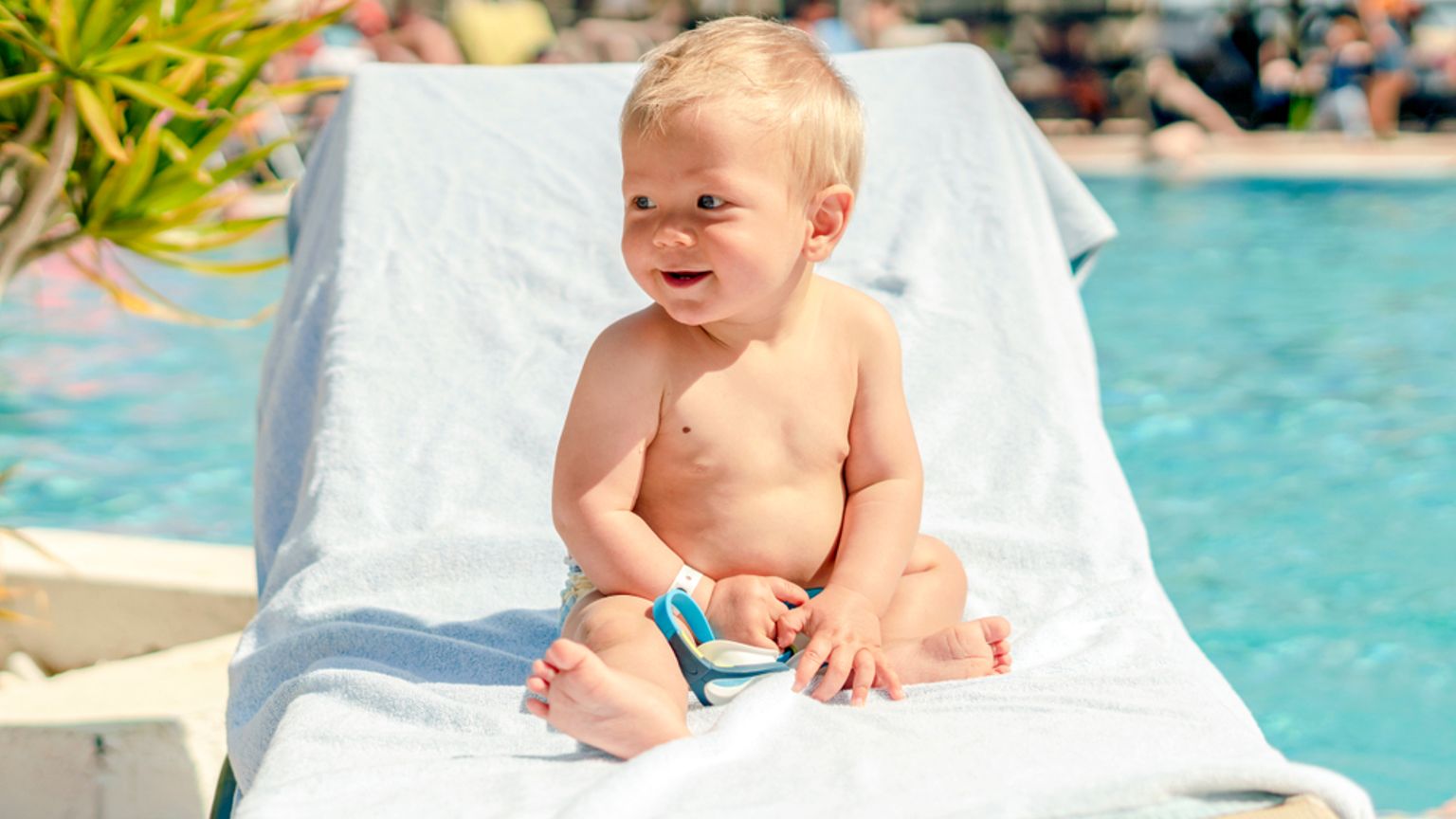 En smilende blond baby i en badebleie sitter på en solseng ved siden av et svømmebasseng.