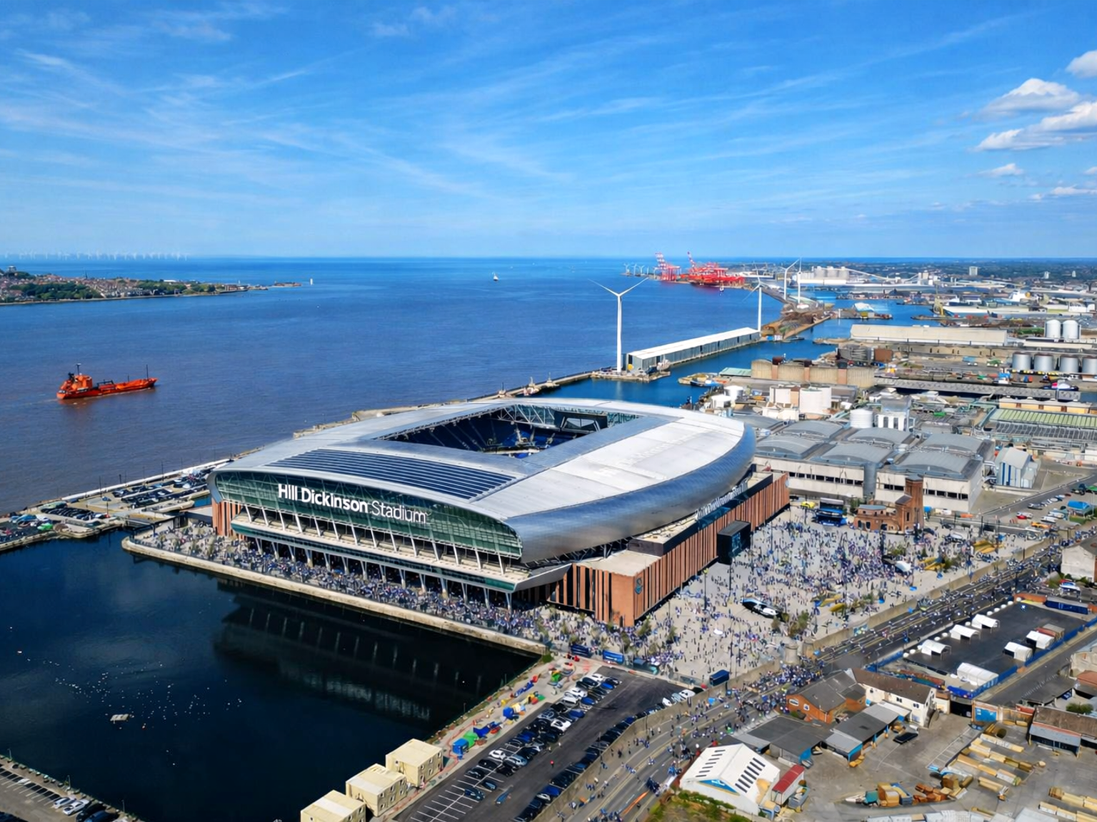 Luchtfoto van het moderne Hill Dickinson Stadium, omringd door menigten, naast water met windturbines en industriële gebouwen op de achtergrond.