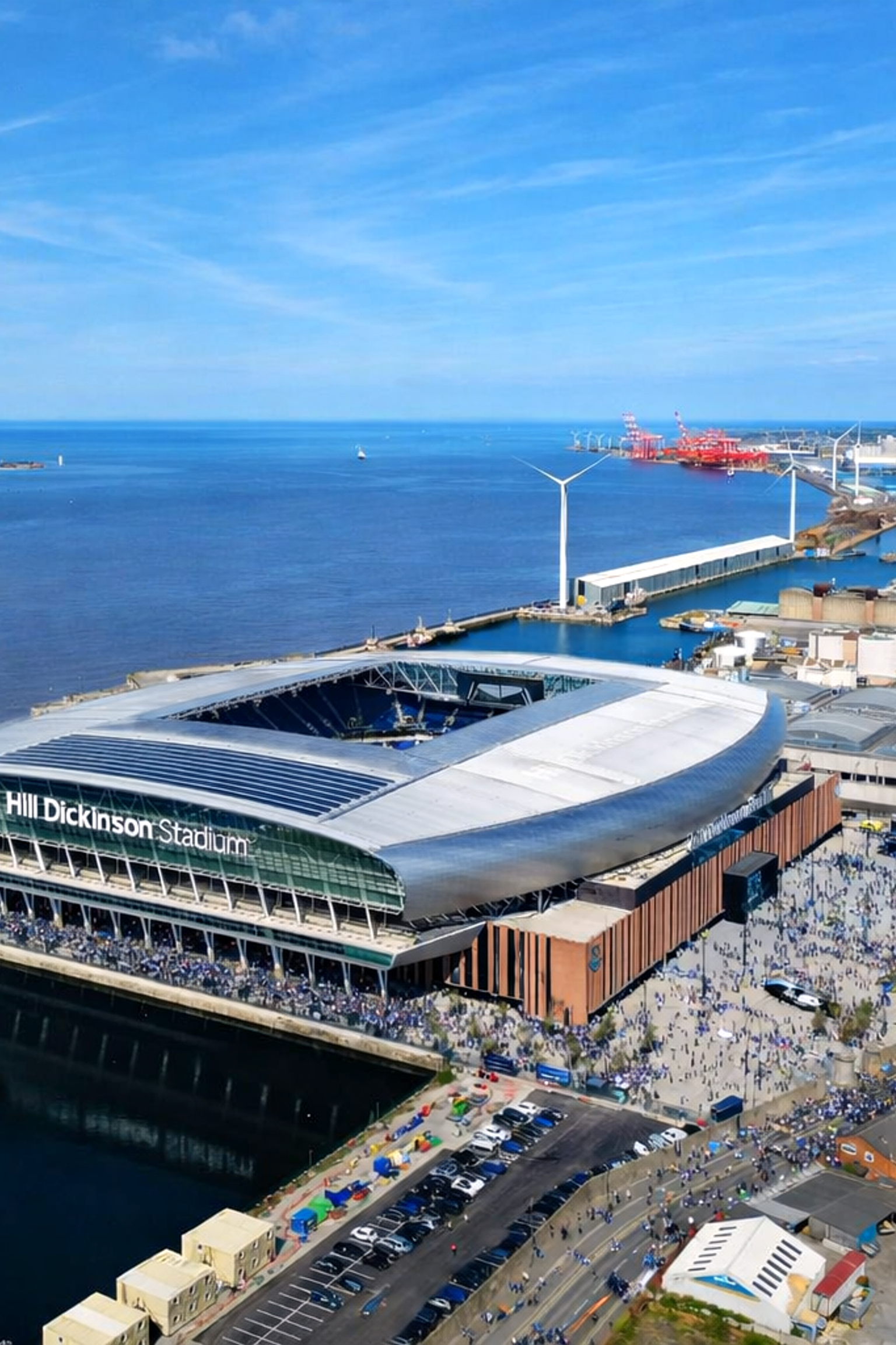 Luchtfoto van het moderne Hill Dickinson Stadium, omringd door menigten, naast water met windturbines en industriële gebouwen op de achtergrond.