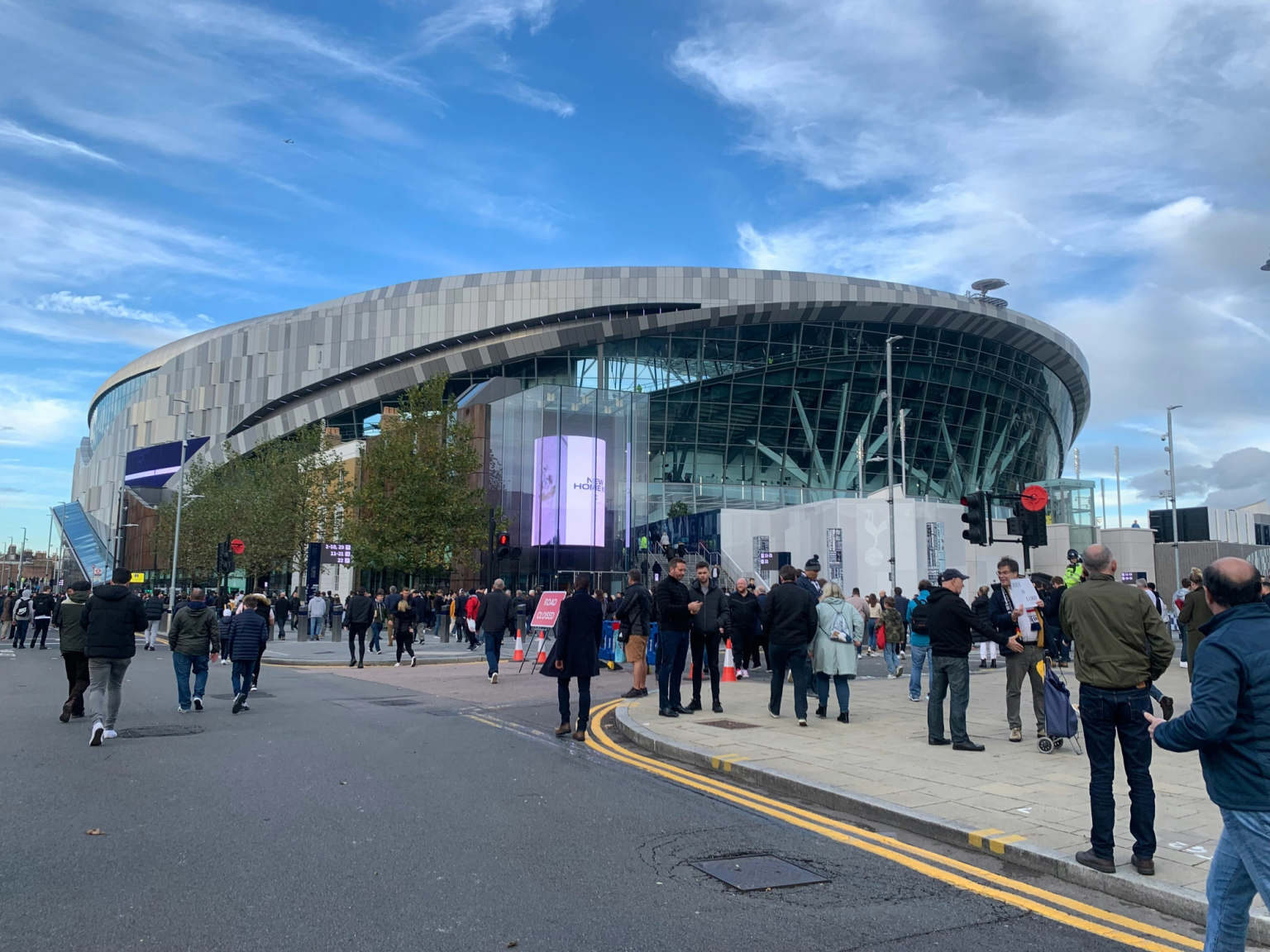 Folkmassor av människor går på en gata och trottoar framför den moderna Tottenham Hotspur Stadium.