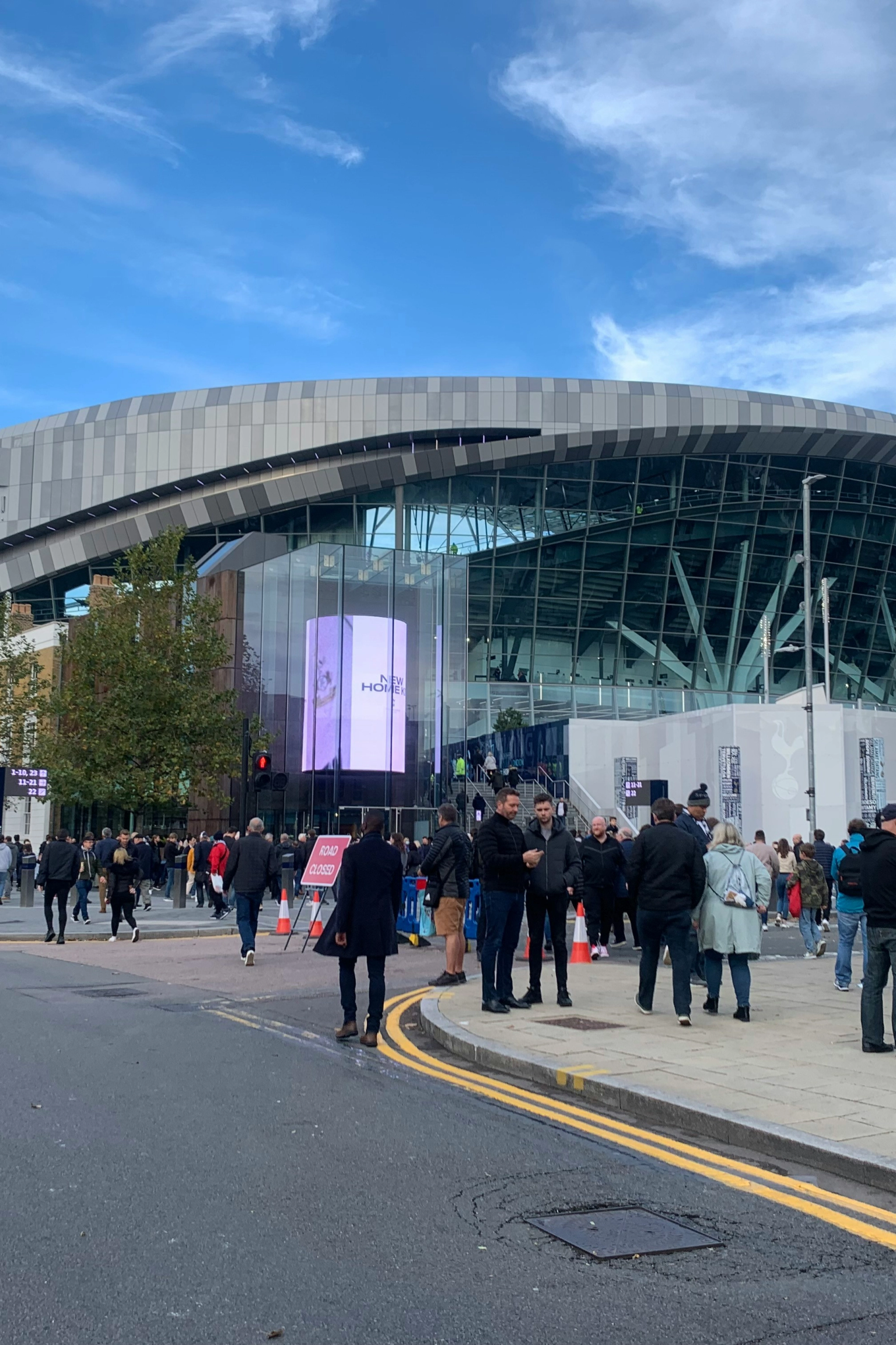 Folkmassor av människor går på en gata och trottoar framför den moderna Tottenham Hotspur Stadium.