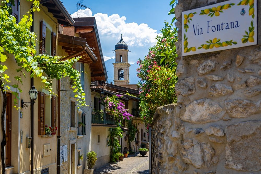 En solrig, smal gade i en italiensk landsby med vinbeklædte bygninger og et klokketårn i det fjerne. Et skilt på en stenmur lyder "Via Fontana" indrammet af citroner. Limone sul Garda, Italien.