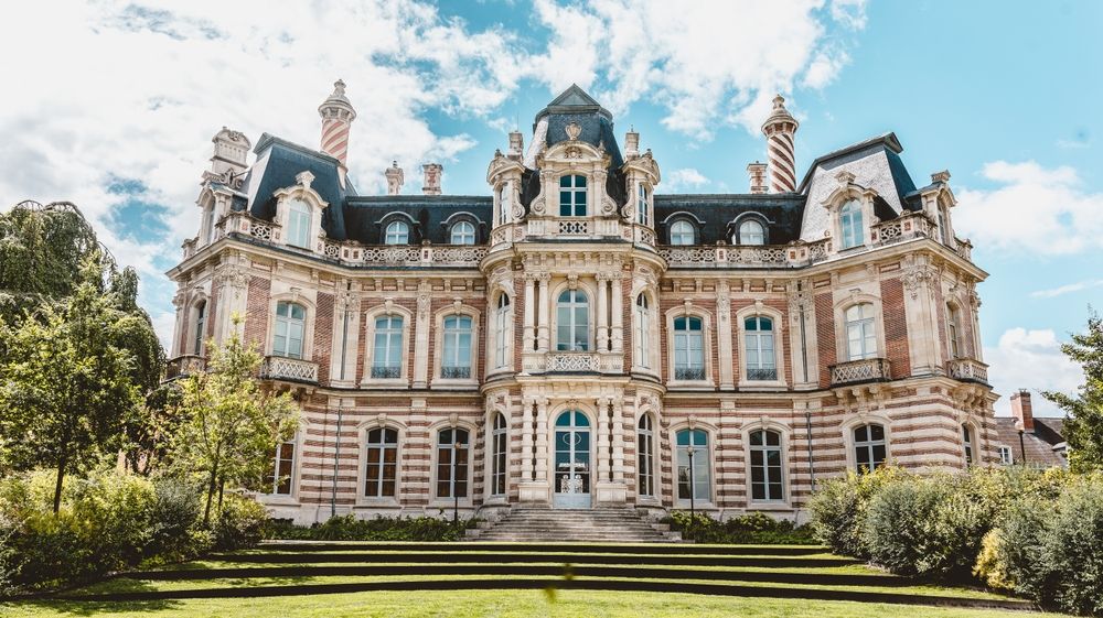 Una mansión ornamentada con una fachada a rayas y tejados oscuros, frente a un césped verde bajo un cielo parcialmente nublado. Château Perrier en la Avenue de Champagne en Épernay.