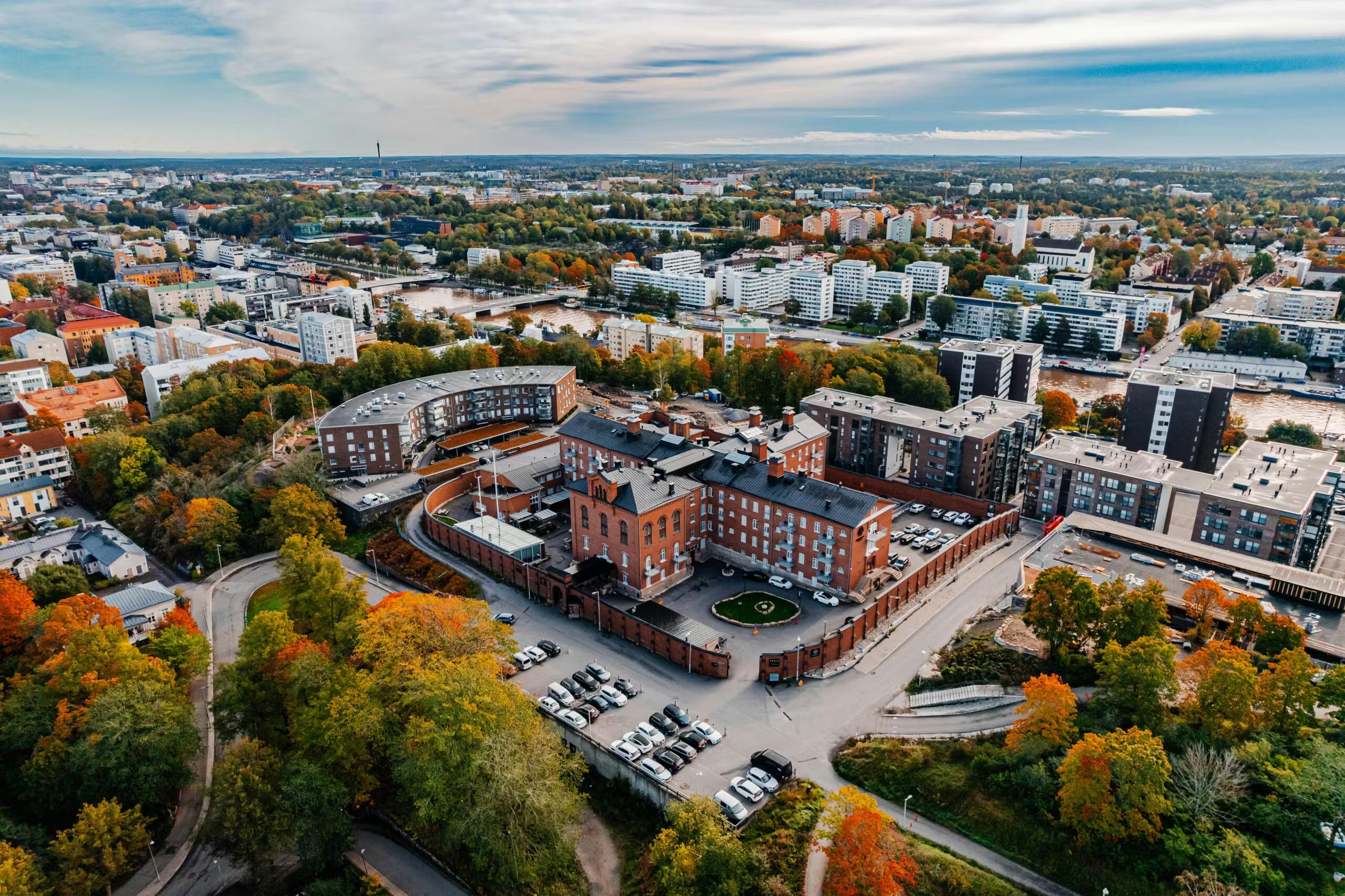 Aerial view of a red brick building complex surrounded by vibrant autumn trees and modern city architecture, with a river winding through.