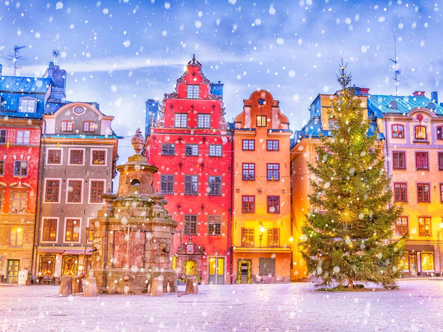 Snö faller på ett festligt torg i Stockholm med färgglada byggnader, en fontän och en upplyst julgran.